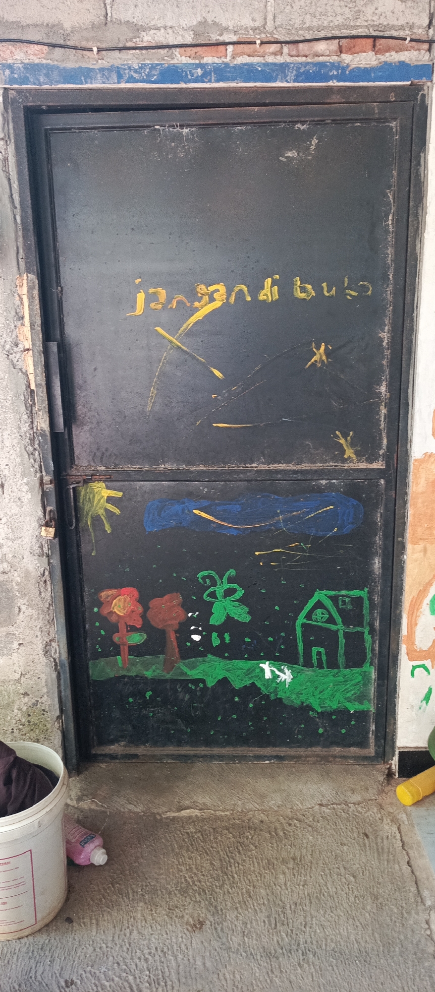 The image shows a black metal door in Arjasa, Indonesia. The top half of the door has yellow writing that appears to say "jangan buba" and some yellow scribbles that look like an X and other abstract shapes. The bottom half of the door is decorated with a child's drawing. The drawing features green grass, two red and orange flowers, a green butterfly, a green house with a circular window, and a small white animal that resembles a dog. Above the grass, there is a blue cloud with yellow streaks and scattered white dots. The door is set into a brick wall, and there is a concrete floor in front of it. To the left of the door, a white bucket with some dark fabric inside and a pink bottle are visible on the floor. A rusted padlock is attached to the door's hinge on the left side.