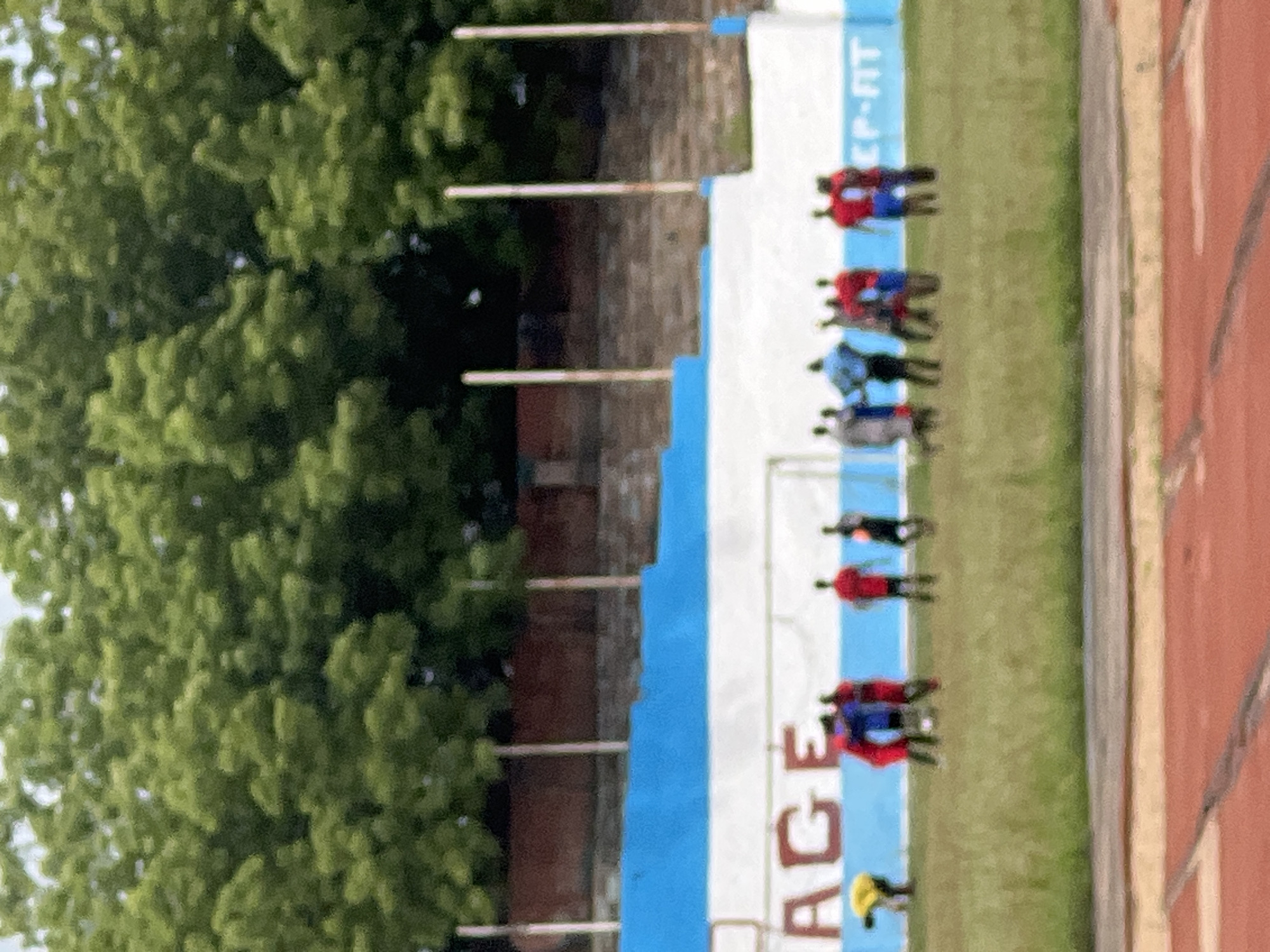 The image shows an outdoor scene with a track and field. A line of people, possibly athletes, are standing on the grass next to a track. The people are wearing various colored athletic wear, including red, blue, and yellow. Behind them is a wall with a blue and white banner that has the words "AGE-FIT" printed on it. In the background are green trees and possibly a brick structure. The location is Unknown, Unknown.