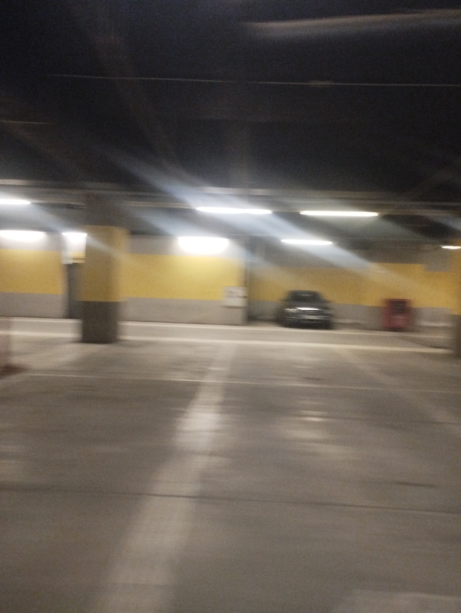 The image shows the interior of a parking garage in an unknown location. The garage has a concrete floor and ceiling, with yellow and gray painted walls. Several fluorescent lights illuminate the space. A dark-colored car is parked further in the background with a red trash can beside it. Support pillars line the garage. The image is slightly blurred suggesting motion during capture. There are no visible people or indications of active interaction.