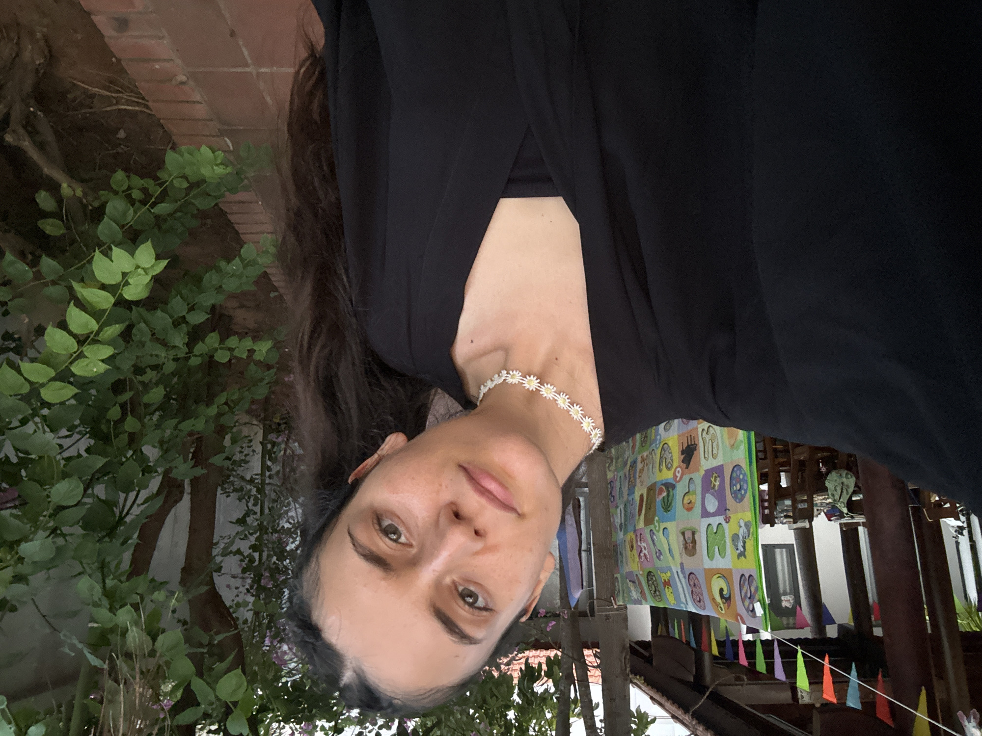 The image presents a portrait of a woman, oriented 90 degrees clockwise, looking directly at the camera with a neutral expression. She has long dark hair, dark eyes, and fair skin, appearing to wear minimal or natural makeup. She is dressed in a black top with a V-neckline and accessorized with a white choker necklace featuring daisy-shaped ornaments.
The setting is an outdoor or semi-outdoor space in Mũi Né, Vietnam. The background on the left (which would be the top when rotated correctly) features lush green foliage and tree trunks, with a brick wall partially visible behind the greenery. On the right (which would be the bottom when rotated correctly), there are indications of a structure, possibly a house or guesthouse, with wooden beams and columns. A prominent colorful fabric banner, likely a children's learning chart, hangs in the background, displaying various illustrated letters, numbers, and shapes. Below this banner, a string of multi-colored triangular flags (bunting) adds a festive or informal touch to the scene.
The lighting is natural and soft, suggesting it might be an overcast day or late afternoon. The overall atmosphere is relaxed and informal. The visible text on the banner consists of fragmented letters and numbers, which are not fully legible for specific words. The primary activity is the woman posing for the photograph.