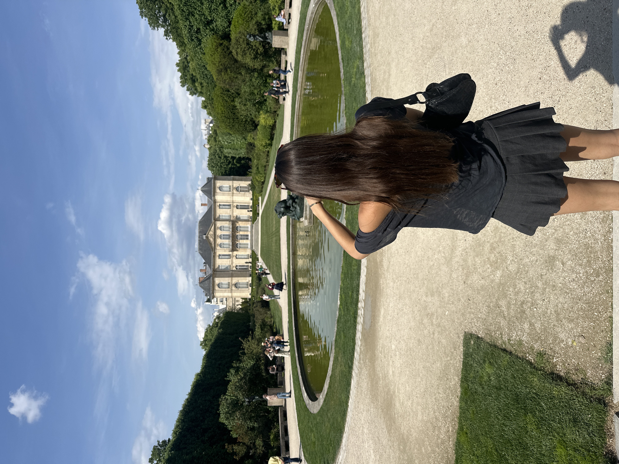 The image captures a person in an all-black outfit standing near a long, oval-shaped reflecting pool. The person has long, dark brown hair and is holding a phone up as if taking a picture of the building across the pool.
The pool is bordered by a narrow strip of grass on both sides, and then a wider pathway where other people are seen walking and sitting. On the other side of the pool is a large, light-colored building with multiple windows, suggesting it's a palace or manor.
The sky is blue with some clouds. The overall setting is a park or garden, with trees and manicured bushes. Based on the architectural style of the building, the image likely shows the Jardin du Palais Royal in Paris, France. A shadow in the foreground right suggests the person is making a heart shape with their hands.