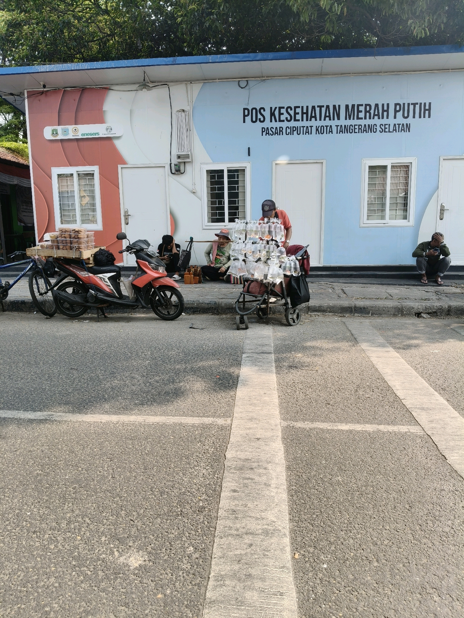 In the city of South Tangerang, Indonesia, on a street with asphalt paving and white road markings, a one-story building with a blue roof is visible. The building's facade is divided into two distinct color schemes: the left side features a reddish-brown hue with a white wavy design, while the right side is painted a light blue. Text on the blue section reads "POS KESEHATAN MERAH PUTIH" above "PASAR CIPUTAT KOTA TANGERANG SELATAN".
In front of the building, on the sidewalk, a man wearing a red shirt and a dark cap is attending to a cart filled with numerous plastic bags, each containing small fish. Adjacent to this, a red motorcycle is parked, with a wooden crate full of bread or pastries secured to its rear. A blue bicycle is positioned to the left of the motorcycle. Further to the right, a person is seated on the ground, looking at their phone, and another person is also seated near the building entrance. Trees are visible behind the building, and the scene appears to be outdoors during the daytime.