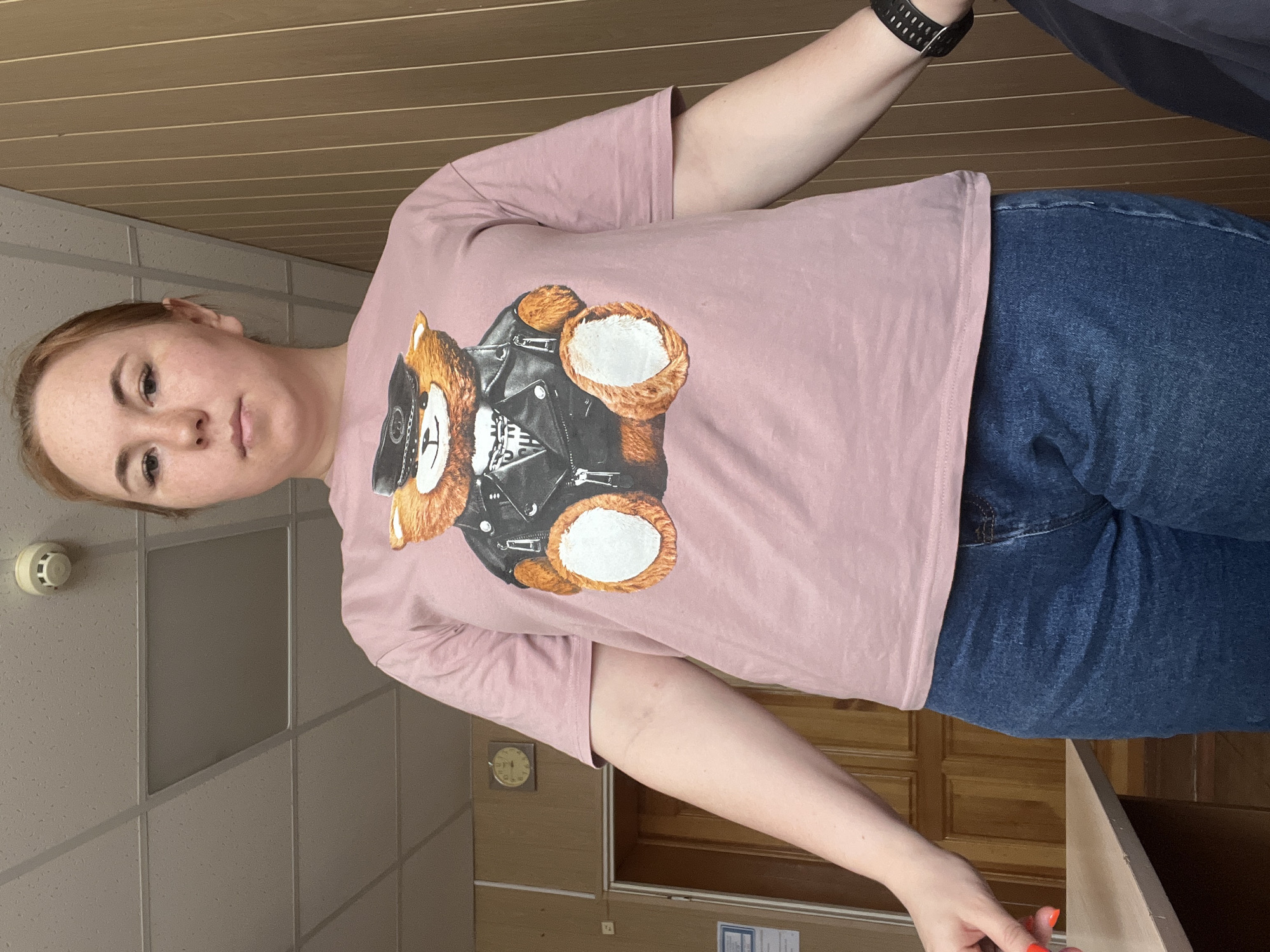 A person wearing a pink T-shirt with a teddy bear in a leather jacket and blue jeans is standing indoors in a room with beige paneling on the ceiling and beige tiled walls. A clock is visible on one of the wooden doors. The location is in the city of Chaikovsky, Russia.