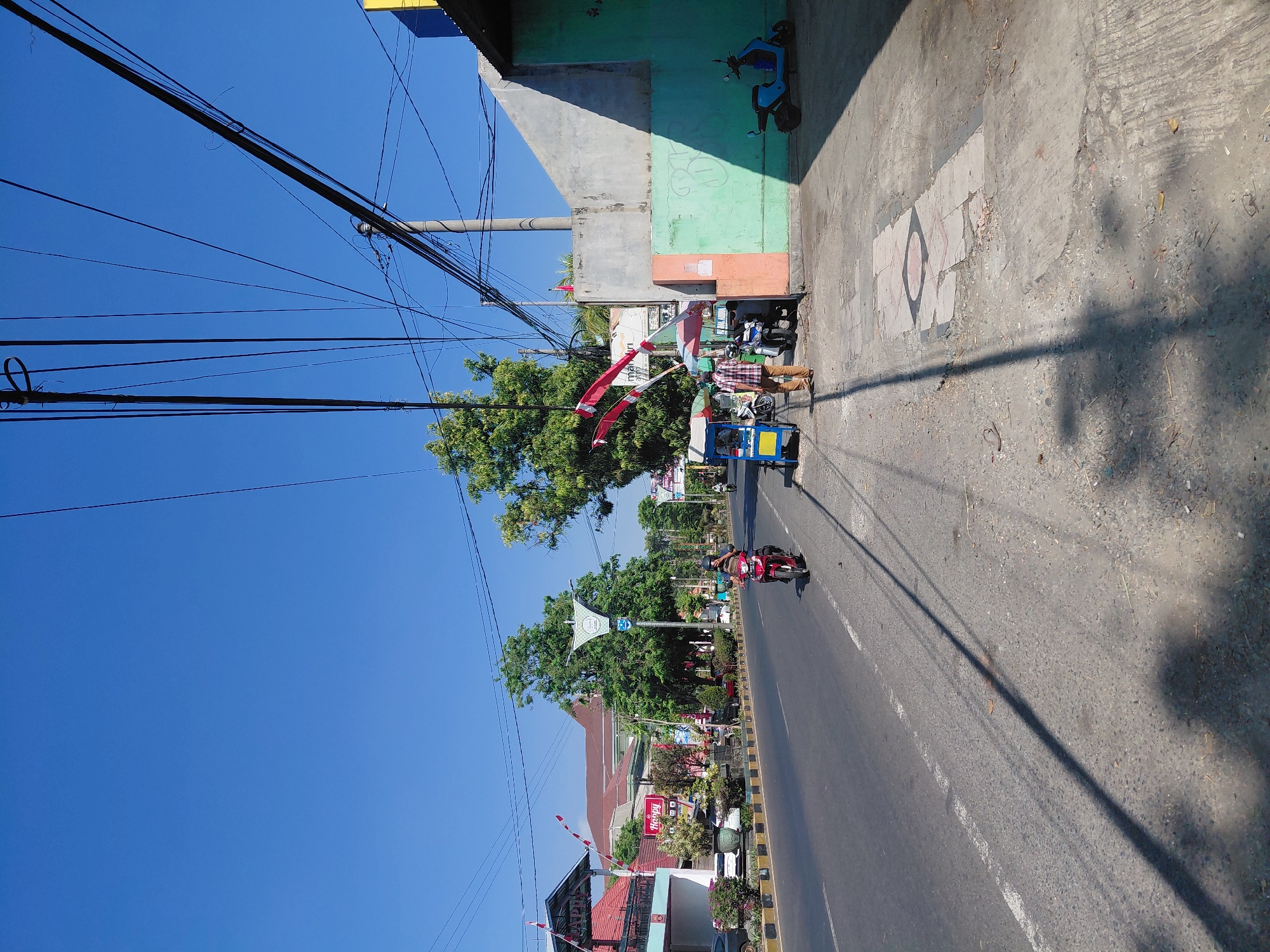 The image depicts a street scene in Semampir, Indonesia, on a clear, sunny day. The perspective is from the side of a road, looking down its length. A two-lane asphalt road dominates the foreground and middle ground. On the left side of the road, there is a sidewalk with various businesses and buildings. One building has a sign that reads "Happy." Further down the road, trees line the sidewalk, and there are more buildings and some parked cars.
On the right side of the road, there is a sidewalk with a concrete wall and a building. The wall is painted with different colors: a light gray section, an orange section, and a light green section. A blue scooter is parked on the sidewalk in front of the green wall. There are power lines strung across the sky, supported by utility poles.
In the middle of the road, a red motorcycle with two occupants is traveling away from the camera. Further ahead, a small blue and yellow cart, possibly a food cart, is parked on the side of the road. Two individuals are standing near the cart. Another red motorcycle is visible further down the road on the left side of the frame. The overall environment appears to be an urban or semi-urban area with moderate traffic.