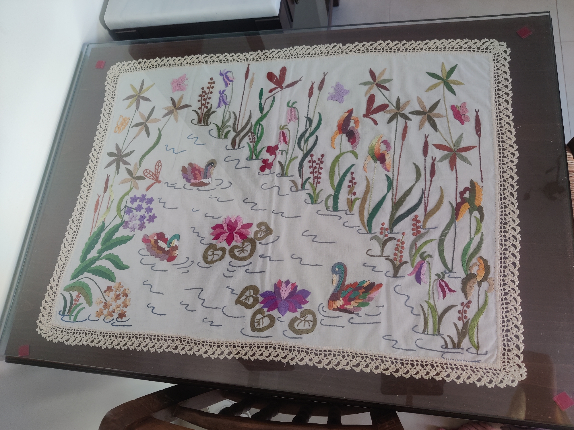 A framed embroidered cloth depicting a pond scene with flora and fauna is displayed on a glass-topped table. The embroidery features colorful flowers, reeds, dragonflies, and birds, possibly swans, in various shades of pink, purple, orange, green, and brown. The cloth has a crocheted lace border. The table itself is dark wood and appears to be in an indoor setting, likely a home, with a white object and a chair visible in the background. The scene is observed from a slightly elevated angle, with the camera capturing the top surface of the table and the embroidered cloth. The location is identified as Katerini Municipality, Greece.