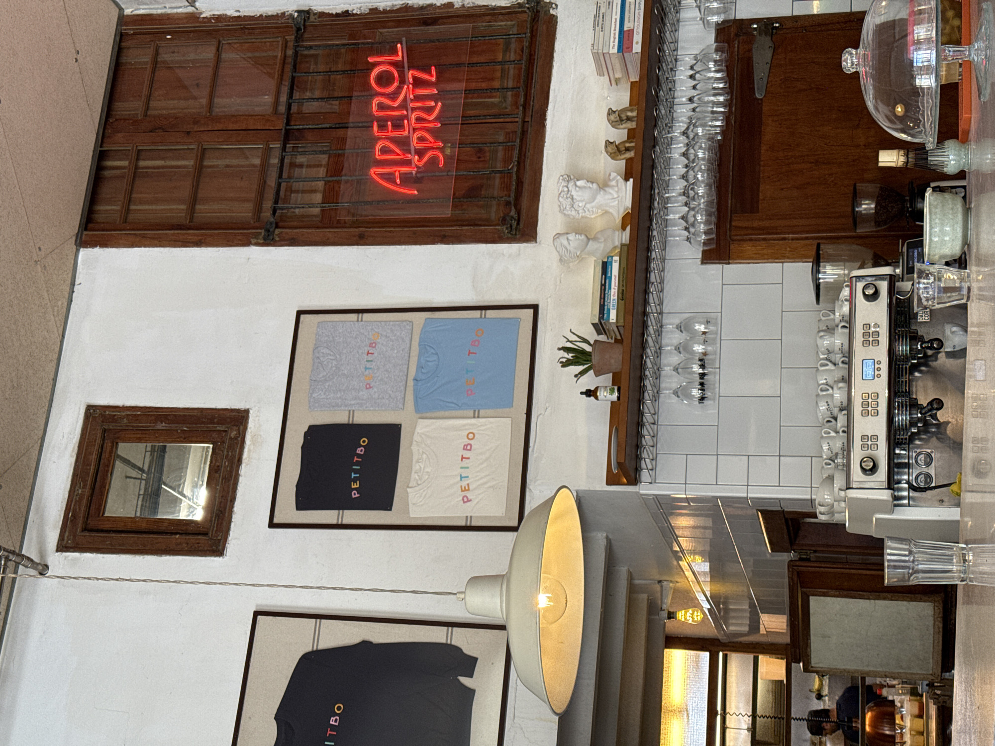 The image shows an interior scene, likely in a shop or cafe, possibly in Barcelona, Spain, due to the "APEROL SPRITZ" neon sign which is common in bars and cafes. The camera is oriented to show a wall display.
On the wall, there are two small, framed rectangular windows, with wooden frames, placed close to the ceiling. A neon sign reading "APEROL SPRITZ" is mounted within one of these window frames. There are also framed artworks featuring t-shirts, with the brand name "PETITBO" visible. A pendant light fixture is hanging down. Shelves are mounted on the walls displaying glasses, books, and decorative busts. A professional-grade coffee machine is visible. A counter or table is in the foreground.
