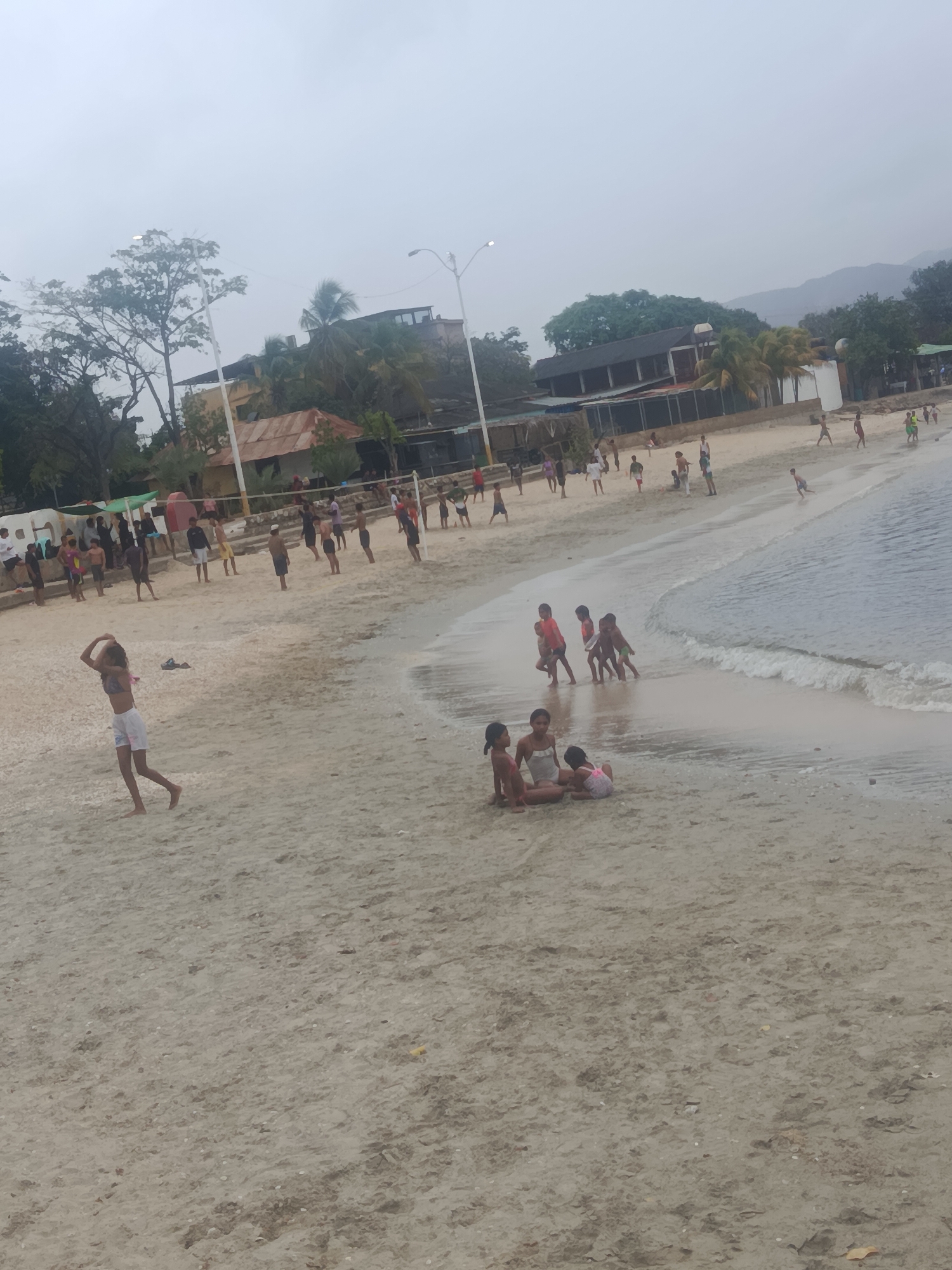 The image shows a beach scene in Juan Griego, Venezuela. A group of children are playing in the shallow water near the shore. In the background, there are buildings and trees. The sky is overcast. There are many people on the beach, some of whom appear to be playing volleyball. The sand is light brown, and the water is a light blue. The overall impression is one of a relaxed, family-friendly beach.