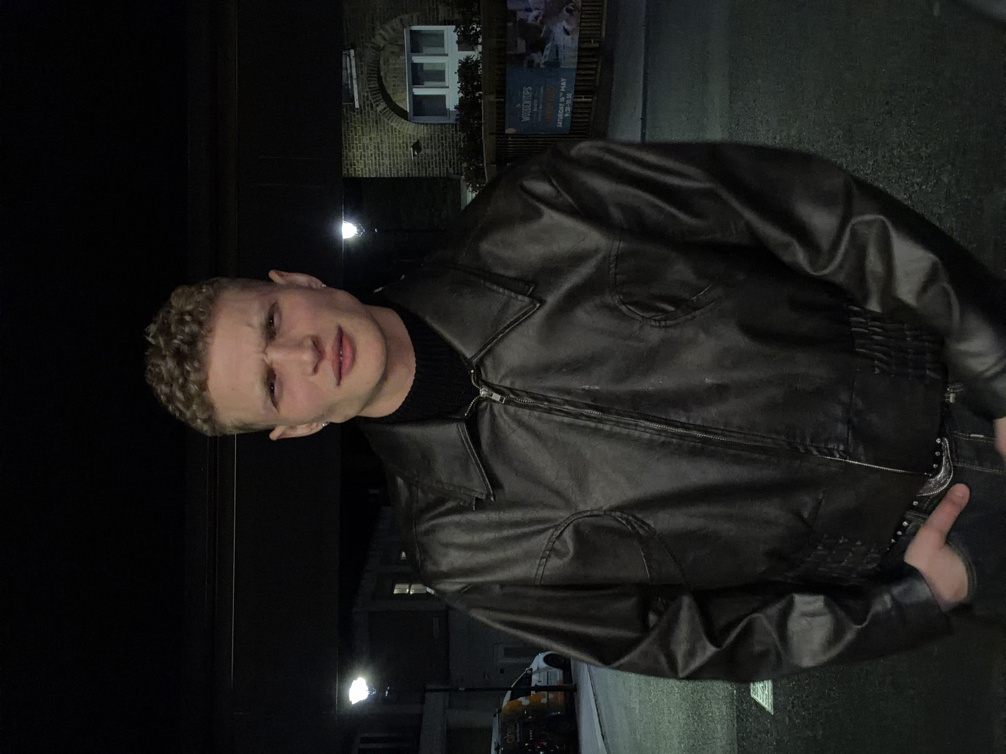 A young adult male with light skin and short, curly blonde hair is seen wearing a black turtleneck sweater and a black leather jacket. He is slightly turned towards the camera, with a neutral expression on his face. The background indicates an urban setting at night, likely in London, United Kingdom, based on architectural details and street signage. Streetlights illuminate the scene, casting shadows and highlighting a building with a window. A sign with the text "WOODENTOPS" is visible. A white van can be seen parked on the street. The image seems to be taken at night on a city street.