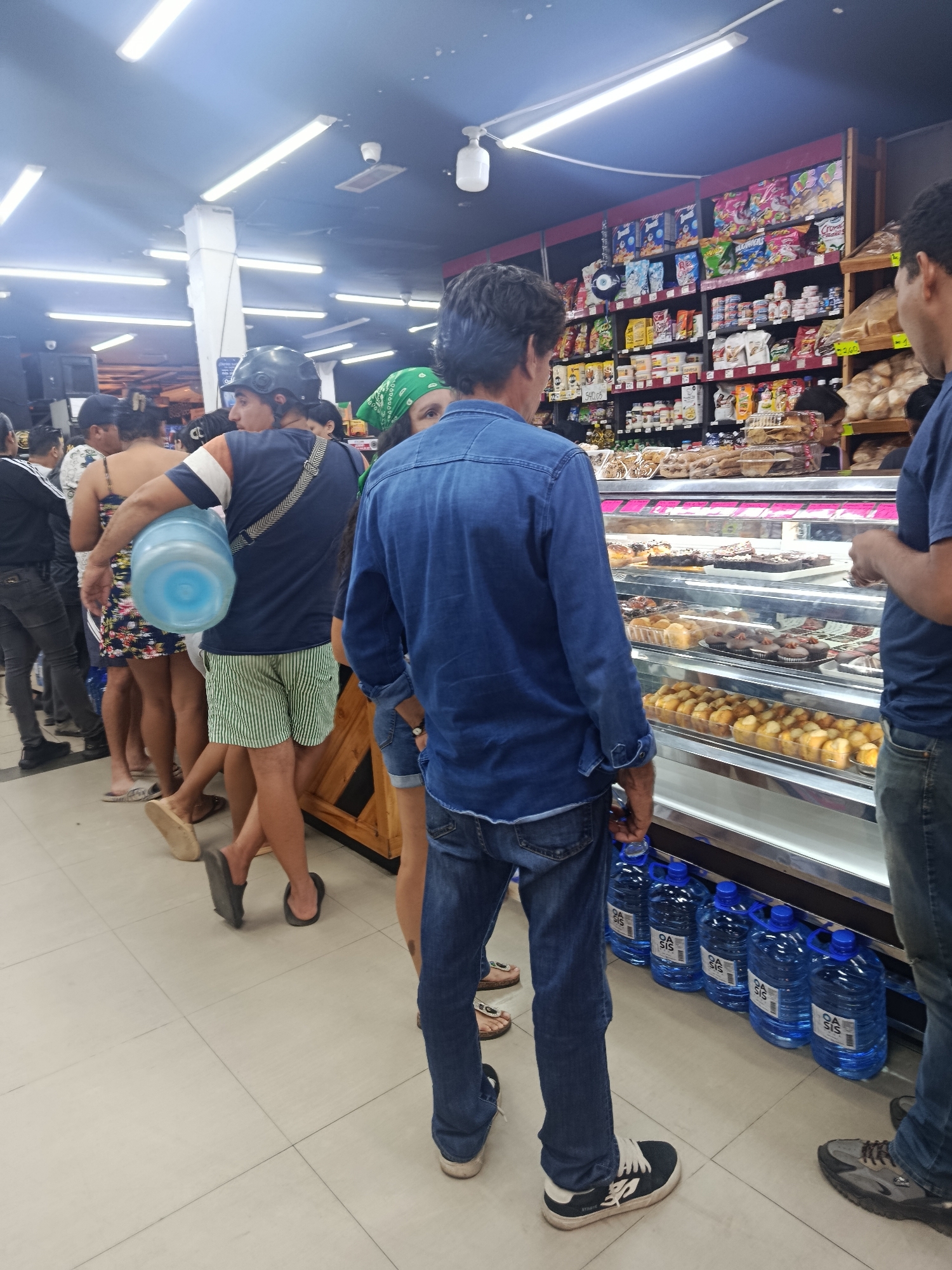 People are lining up at what appears to be a bakery or grocery store in Altagracia, Venezuela. The lineup is on the left side of the frame. The store has a well-lit interior with fluorescent lighting and shelves stocked with packaged foods. A refrigerated display case filled with pastries is visible. There are multiple large bottles of water with blue caps on the floor by the display case. One man in the line is wearing a helmet and carrying a large water bottle.