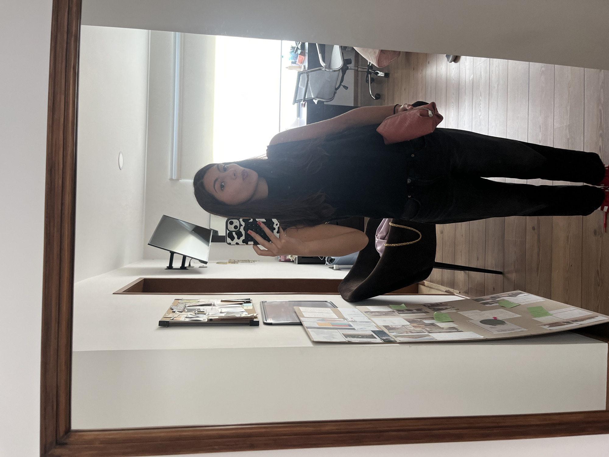 A woman is taking a selfie in a mirror. The mirror has a wooden frame. The woman is wearing a dark top and dark jeans. She is holding a cell phone with a patterned case. A pink pouch is on her left shoulder. In the background, there's a room with a wooden floor. A white desk is in front of the mirror, and on the desk are various papers, photos, and a laptop screen. A chair is visible near the desk. The location is indeterminate.