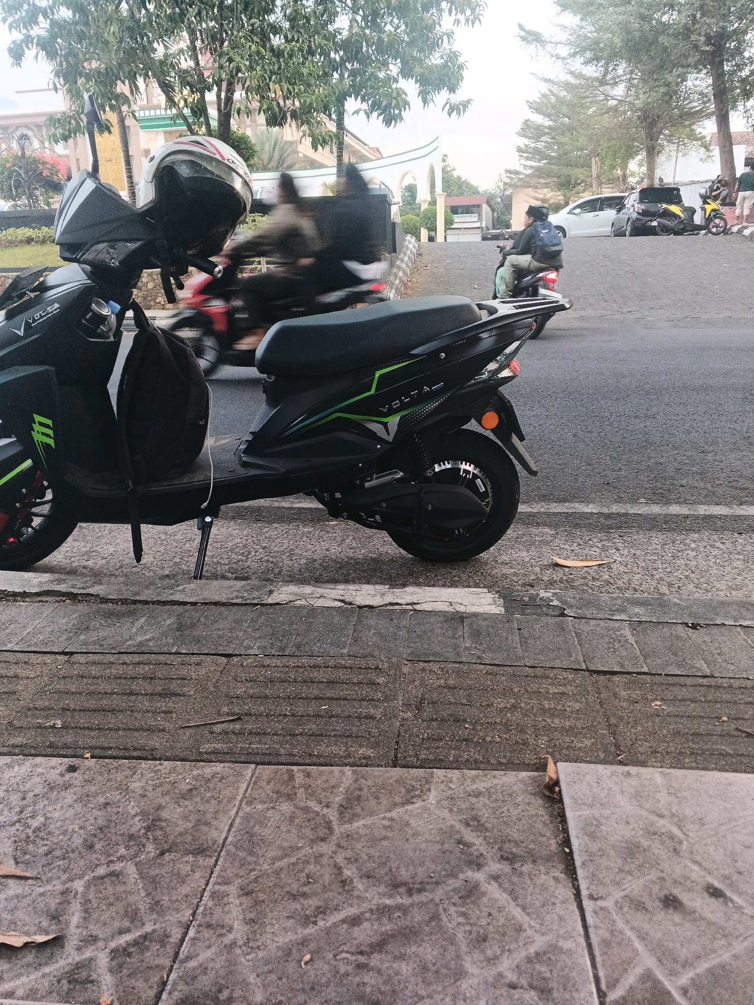 The image depicts a street scene in Sidomulyo, Indonesia, with a black electric scooter parked on the side of the road. The scooter has green accents and a helmet resting on its handlebars. A backpack is also visible on the scooter. In the background, other vehicles, including motorcycles and cars, are present on the road. Trees and a building with a distinctive architectural style are visible further in the distance. The pavement in the foreground consists of textured gray tiles. The overall lighting suggests it is daytime.