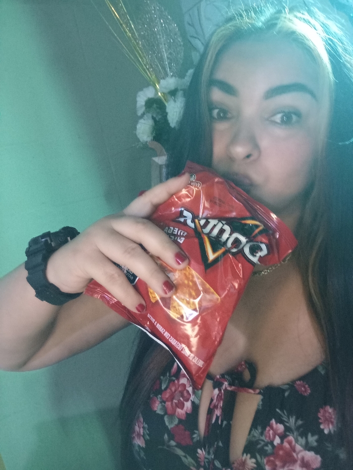 A woman with highlighted hair holds a red bag of Doritos close to her face. She has red nail polish on and wears a black watch. A floral print dress is visible. In the background, there's a light green wall, a decorative floral piece, and shimmering gold tinsel.