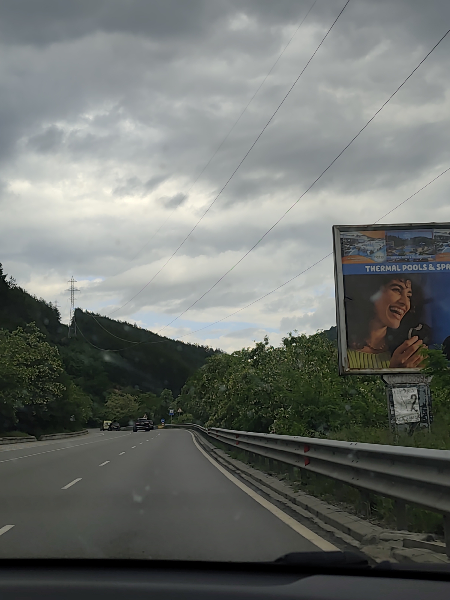 The image is taken from a moving vehicle on a road near Gradevo, Bulgaria. The sky is overcast. A billboard advertising "THERMAL POOLS & SPA" with an image of a smiling woman is visible on the right side of the road. The road is bordered by a metal guardrail. In the distance, there are green hills covered with trees. An electrical transmission tower is visible on the hill. Several vehicles are further up the road, including one possibly stopped on the side. There are power lines visible in the sky.