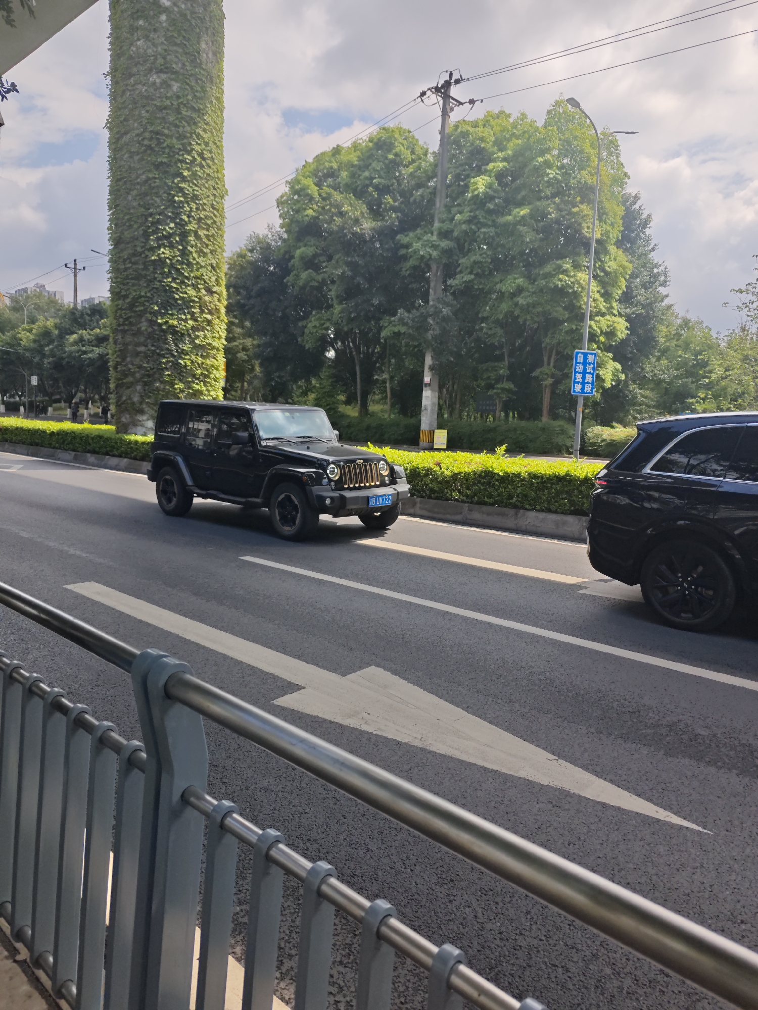 The image shows a street scene in Chongqing High-tech Zone, China. A black Jeep Wrangler is driving on a road with a white directional arrow painted on the asphalt. A black SUV is positioned to the right of the Jeep. A tall, cylindrical structure covered in green vines stands adjacent to the road. A blue sign with Chinese characters is visible near the curb. A metal guardrail runs along the edge of the road. Trees and a street light are visible in the background. The sky is partly cloudy.
