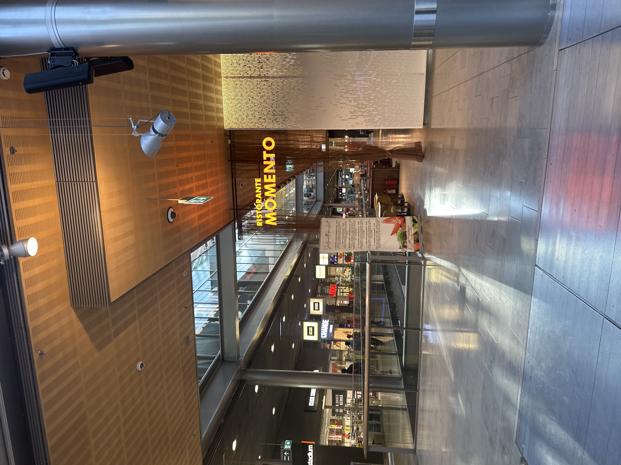 The image depicts the interior of a modern shopping mall. The camera is angled upwards, giving a view of a multi-level structure with shops and restaurants. In the foreground, a large metallic pillar dominates the left side, with lighting fixtures and what appear to be security cameras mounted on the ceiling above.
The dominant visual element is the illuminated signage for "RISTORANTE MOMENTO," a restaurant with a distinctive yellow, glowing logo. The restaurant is draped with sheer brown fabric, creating an inviting entryway. Further into the mall, various shopfronts are visible, including "CHANGE" and "CLK SHOES."
Sunlight streams through the upper levels of the mall, casting distinct geometric patterns on the tiled floor, suggesting it is daytime. The architecture features a combination of wood-paneled walls with perforated patterns and sleek, modern glass and metal structures. Some people can be seen sparsely distributed throughout the mall, suggesting a moderate level of foot traffic.
A menu board is visible in the lower right portion of the image, displaying food items in Finnish, such as "CHEVRE KANACAESARSA" and "LOHI-CAESARSALAATTI." Another sign indicates "stadium." The overall impression is that of a well-lit, contemporary commercial space.