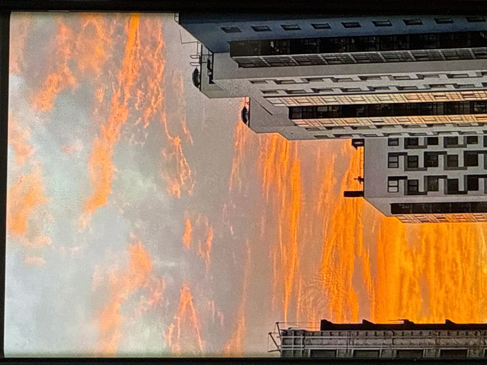 The image captures a vibrant sunset over a cityscape. The sky is ablaze with hues of orange and pink, with wispy clouds streaked across the horizon. In the foreground, modern residential buildings rise into the sky, their glass windows reflecting the warm light of the setting sun. The architectural styles of the buildings are varied, suggesting a mix of contemporary design. One building on the right is tall and slender, with a blue facade and numerous windows. To its left, another building is lighter in color, featuring a more angular design. On the far left, a portion of another building is visible, appearing darker and taller. The scene evokes a sense of urban tranquility as day transitions into night.