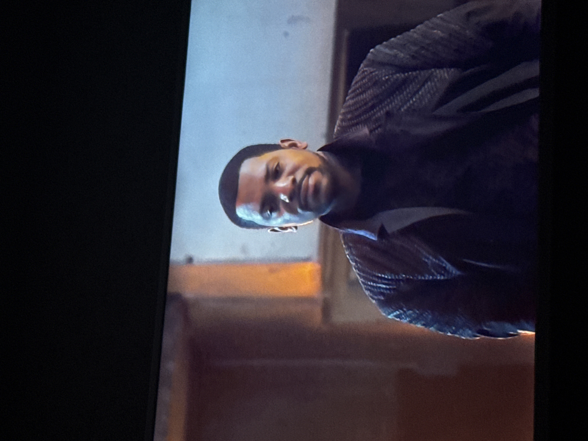 The image shows a man with short dark hair, light brown skin, and dark eyes. He is wearing a dark jacket with a textured pattern on the shoulders. The background appears to be an interior setting with blurred shapes and warm lighting. The image is slightly rotated counter-clockwise. It is not possible to determine the location of Mytishchi, Russia, based solely on the visual content of this image.
