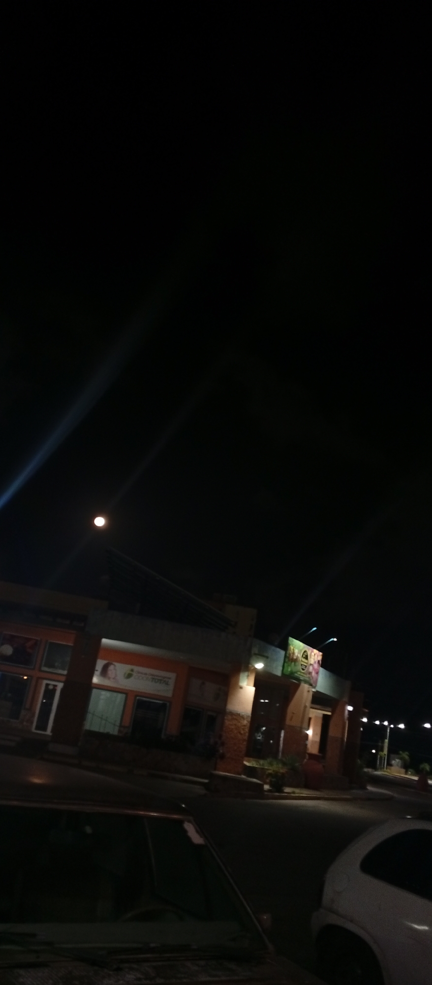 A low-angle shot taken at night features a street in Juan Griego, Venezuela. A building with several storefronts is visible, illuminated by streetlights. The name of the building is unknown. Two cars, one dark-colored and one white, are parked on the street. The sky is dark. There are no visible people.