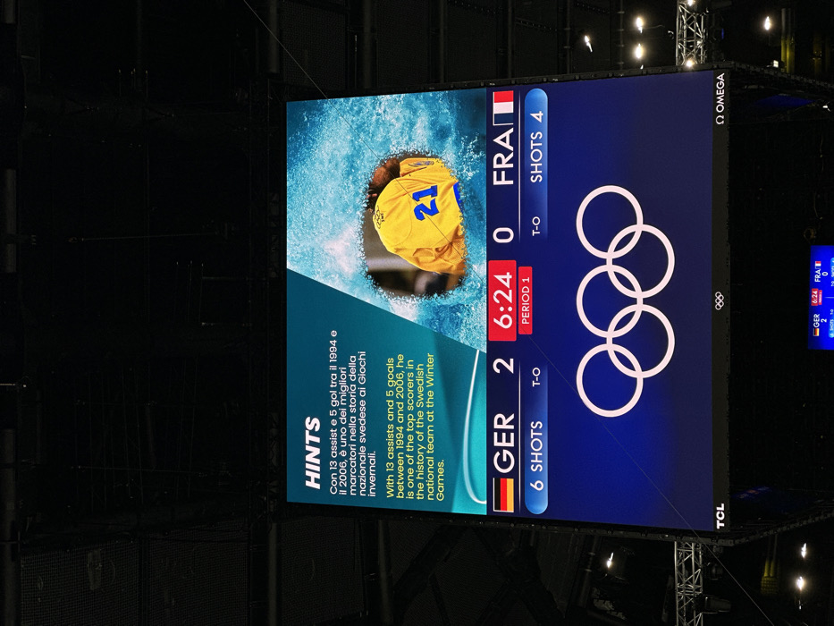 The image displays a large digital scoreboard prominently positioned within a dark indoor arena, consistent with a sports venue in Milan, Italy, likely associated with the Milano Cortina 2026 Winter Olympics given the context of the Olympic rings and the smaller screen below.
The upper section of the screen features a "HINTS" box. This box contains text in both Italian and English, describing a player with 13 assists and 5 goals between 1994 and 2006, identified as one of the top scorers for the Swedish national team at the Winter Games. To the right, a circular image depicts the back of a person wearing a yellow jersey with the number "21" and a "SWE" logo, surrounded by a light blue, splash-like graphic.
The middle section of the scoreboard shows a live game score: "GER" (Germany) with 2 points, and "FRA" (France) with 0 points. A timer indicates "6:24" remaining in "PERIOD 1". Below this, Germany has "6 SHOTS" and France has "4 SHOTS".
The lower section of the screen features the iconic white Olympic rings logo centered on a dark blue background. The bottom left corner of the entire display shows the "TCL" logo, while the bottom right corner displays the "OMEGA" logo.
The surrounding environment is dark, revealing the structural elements of the arena's ceiling, including metal trusses, cables, and various overhead lighting fixtures, indicating an indoor setting. A smaller, identical digital scoreboard is visible further below the main display.