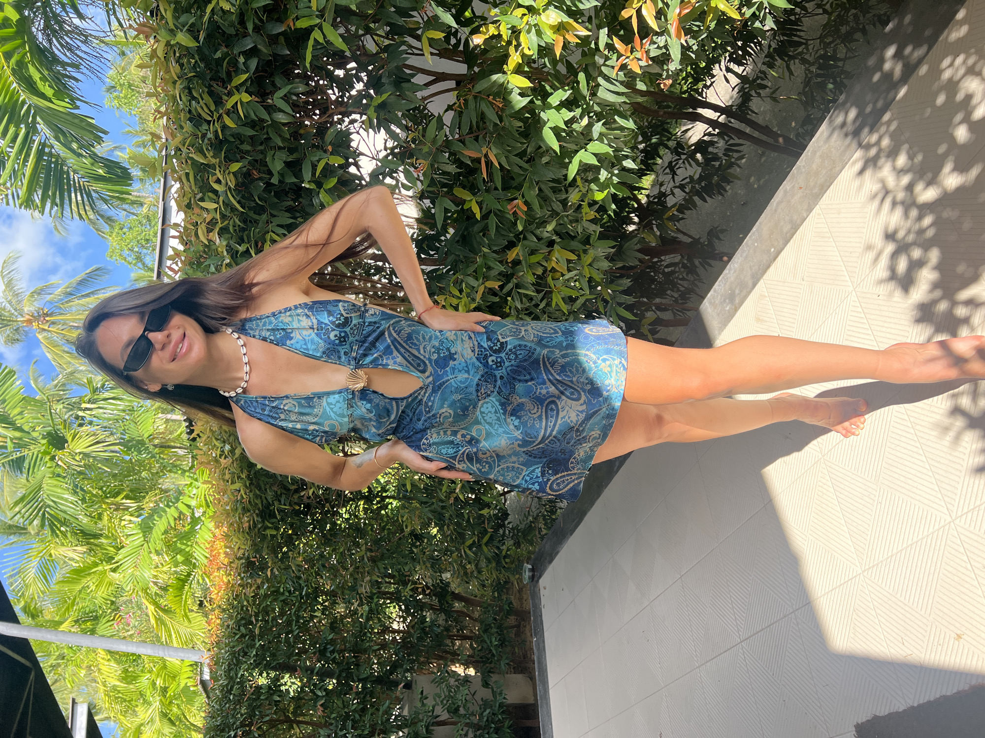 A woman with long brown hair and sunglasses stands outdoors, looking at the camera. She is wearing a blue patterned halter dress with a cut-out detail at the chest, adorned with a shell-shaped clasp. She has a shell necklace and a thin gold bracelet. The background features lush green foliage and palm trees under a bright, sunny sky. The woman is standing on a tiled patio, with a shadow cast across the tiles. The setting appears to be a tropical environment, possibly in the vicinity of Ban Bang Rak, Thailand.