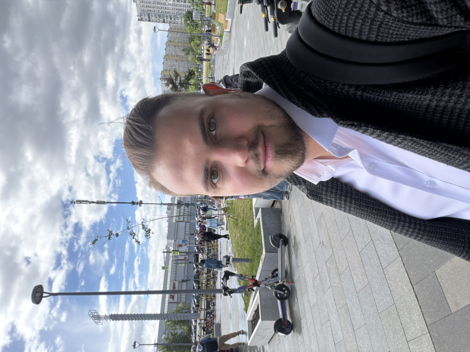 A medium-shot selfie of a person with short brown hair, a light beard, and green eyes, looking at the camera. They are wearing a white collared shirt under a dark gray patterned sweater or jacket, and what appears to be a black backpack.
The person is outdoors in a city environment. The background includes a wide walkway made of gray rectangular pavers, with a section of grass on the left side. Behind the grass, there are buildings, light posts, and other pedestrians. In the immediate background to the right are multiple parked electric scooters. The sky above is blue with scattered clouds.