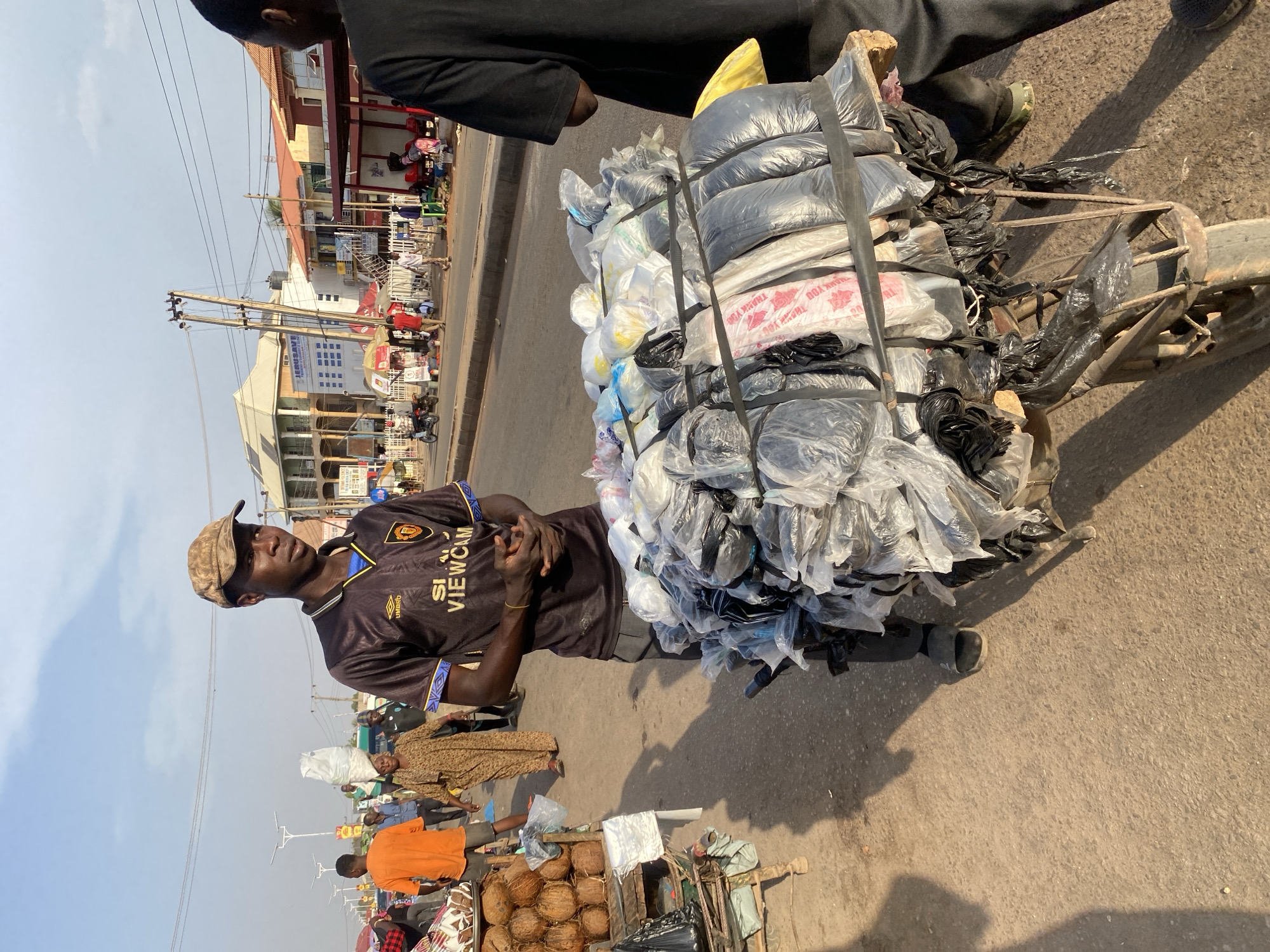 An outdoor scene in Sabon Tasha, Nigeria, depicts a bustling street environment during daylight hours. A young adult male, of dark complexion, stands centrally, wearing a camouflage baseball cap, a dark football jersey with "UMBRO" and "S1 VIEWCAM" branding, grey trousers, and dark sandals. He has a gold-colored bracelet on his right wrist and his hands are clasped in front. He stands beside a modified, old metal cart or bicycle frame, heavily laden with bundles of plastic bags, primarily black and clear, some displaying "THANK YOU" text. The bundles are secured with black straps.
To the immediate right, the back and right arm of another individual wearing a black long-sleeved garment are partially visible. In the midground, to the left of the central figure, a male in an orange t-shirt stands near a stall displaying coconuts. Further back, a woman in a patterned brown dress walks with a white bundle balanced on her head. Numerous other pedestrians and market activities populate the background.
The street is paved, and shadows are cast from the left, indicating the sun's position. Overhead utility poles and multiple power lines traverse the upper portion of the scene. Buildings line the street, including a multi-story structure with "JERUSAVE" visible on its facade. Motorcycles are parked or moving on the far side of the street.