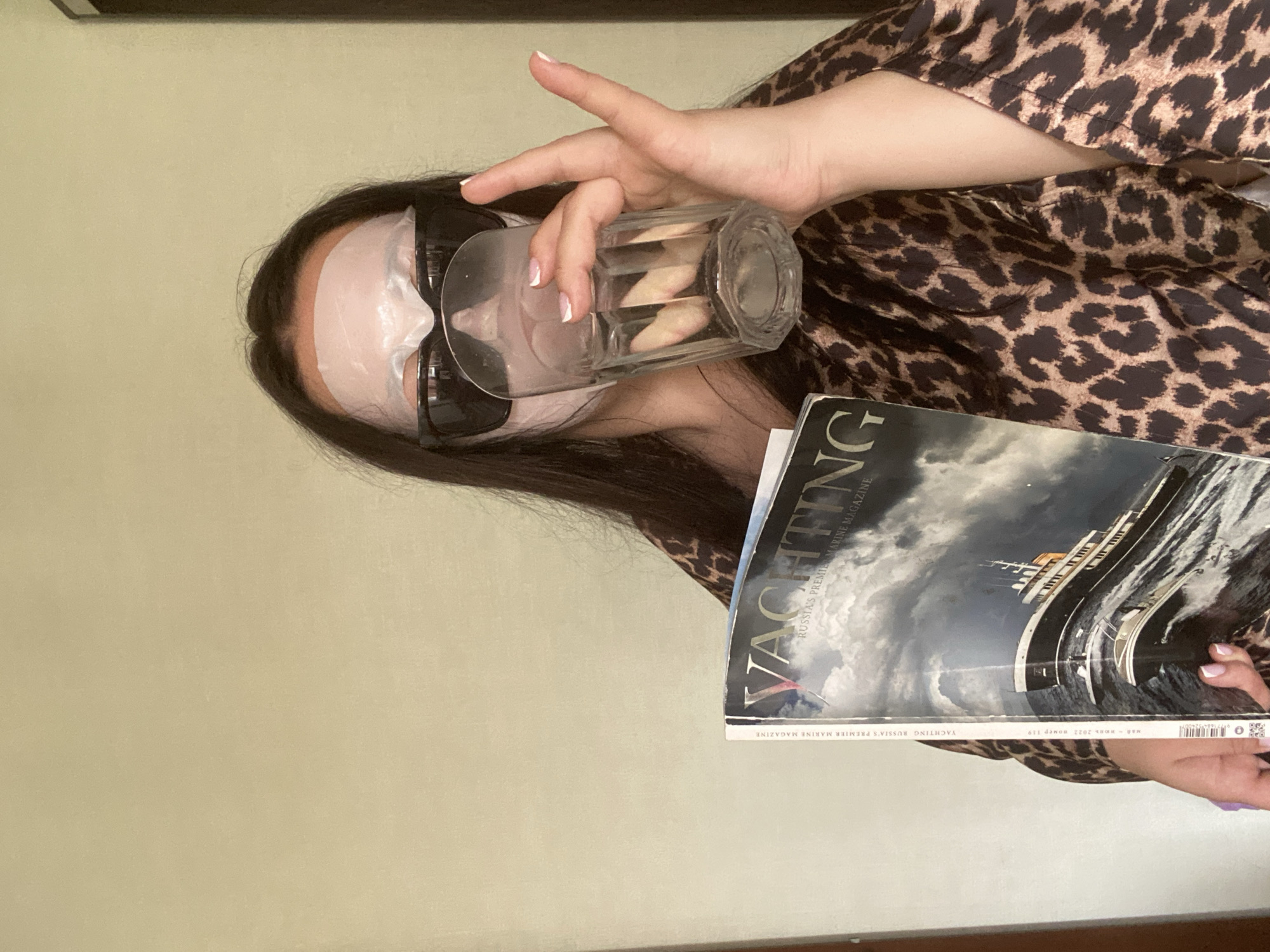 A person wearing a face mask and sunglasses is holding a glass of water and a magazine. The person is wearing a leopard print robe. The magazine is titled "YACHTING RUSSIA'S PREMIER MARINE MAGAZINE" and has "2022 HOMEP 119" printed on the spine. The magazine's cover features an image of a large yacht at sea. The environment consists of a plain beige wall. There is no indication of the city of Sochi, Russia in the image.