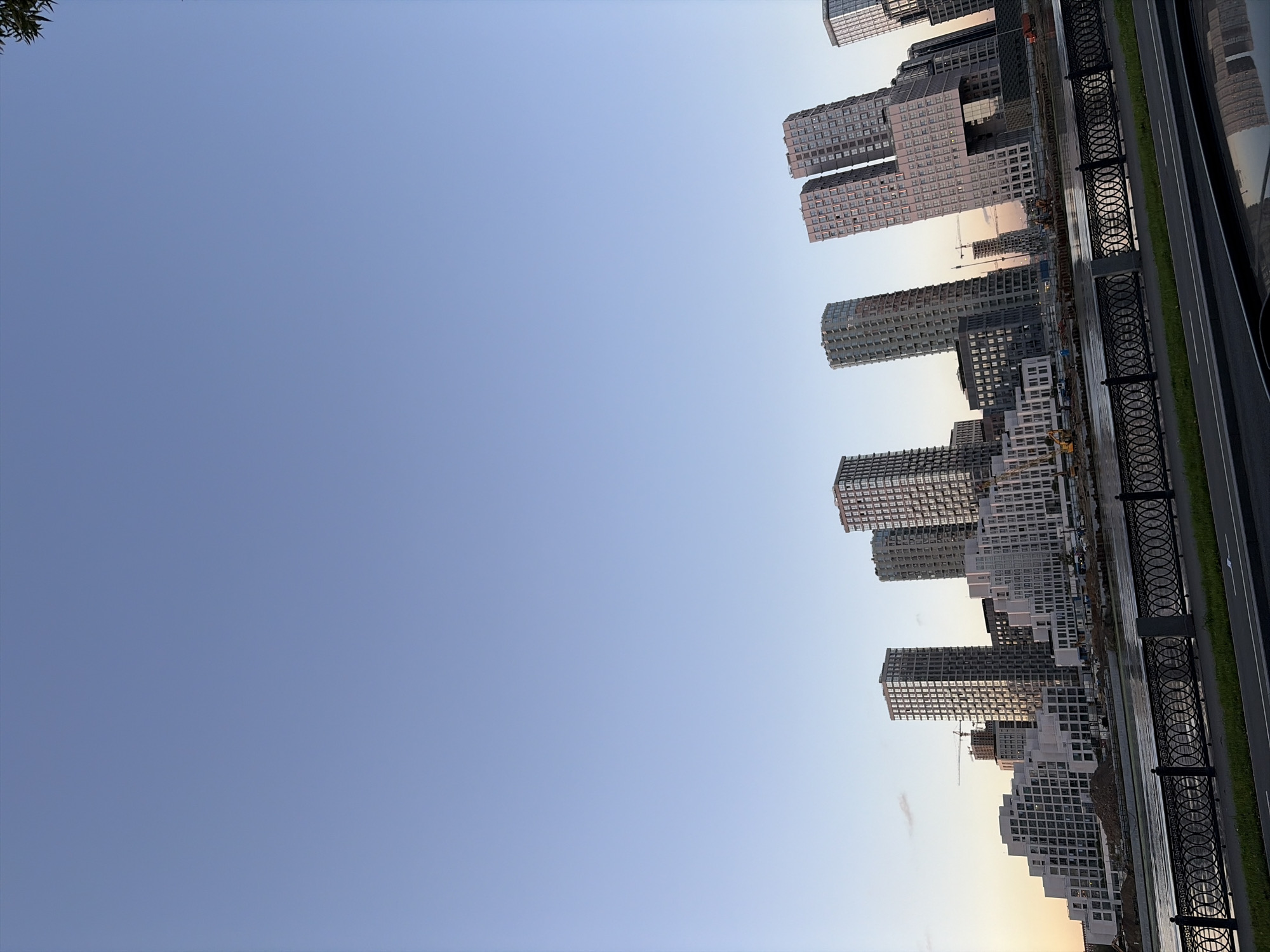 The image shows a view of the Moscow City skyline and residential buildings along the Moskva River. The buildings are modern skyscrapers and a complex of white, stepped apartment buildings. Construction cranes are visible in the background, indicating ongoing development. The river is in the foreground with a decorative metal fence along its bank. The sky is a clear, pale blue with a hint of sunset colors on the left horizon. No people are visible in the image. The overall scene depicts a contemporary urban landscape during what appears to be late afternoon or early evening.