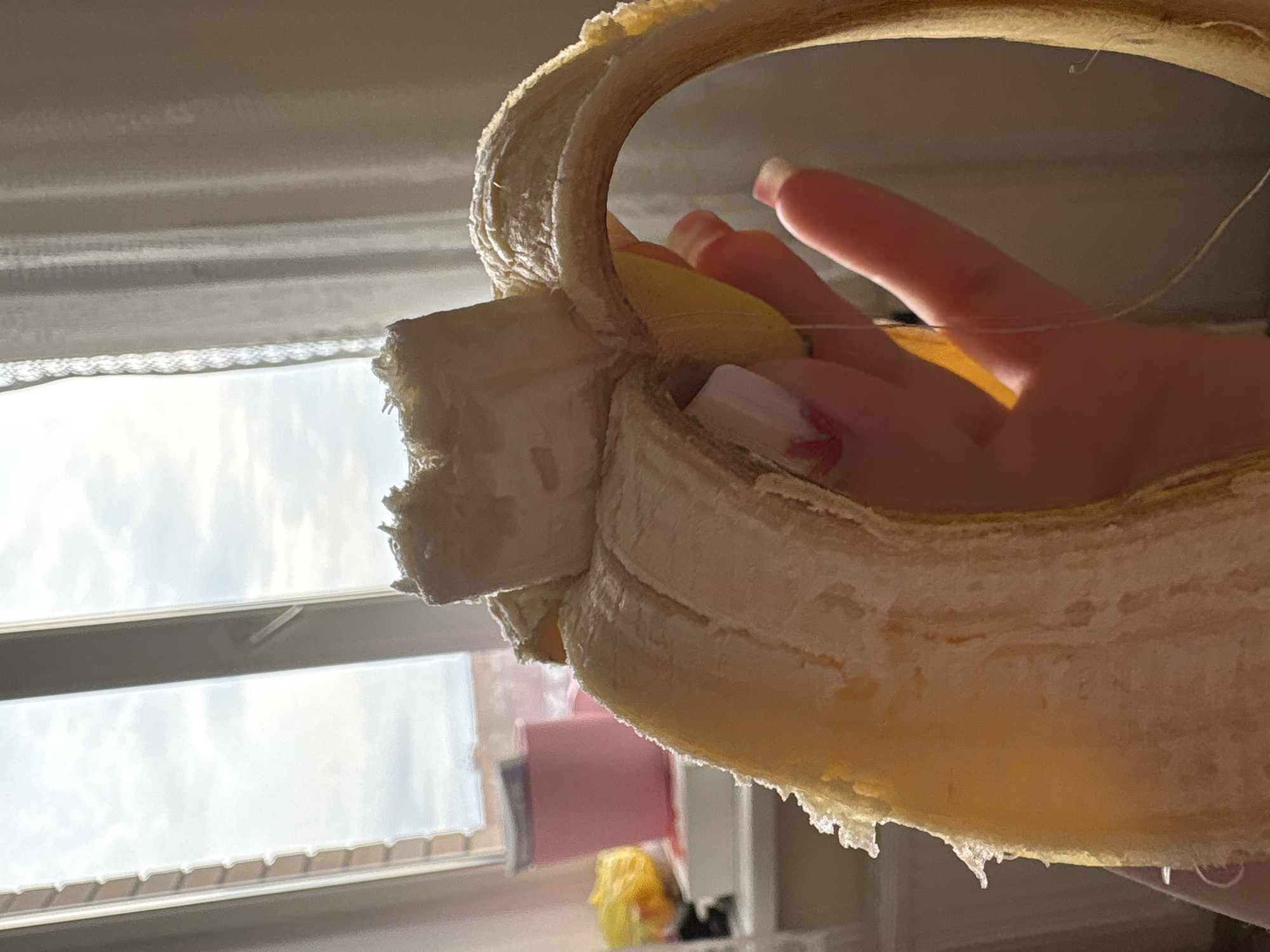 A hand holds a partially peeled banana. The banana peel is arched back. The background features a window with a view of a sky with clouds. Other details from the location are indiscernible. Izhevsk, Russia, is not discernible.