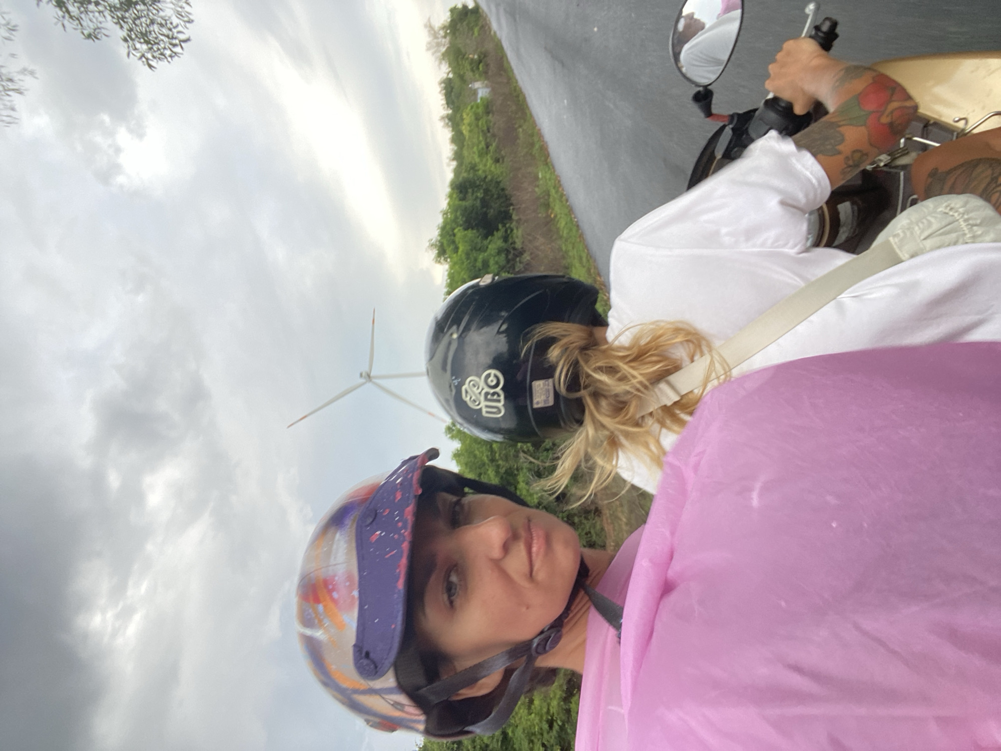Two women are riding on a motorcycle on a paved road, with a scenic view of green hills and a wind turbine in the background. The woman in the foreground is wearing a colorful helmet and a pink poncho, looking directly at the camera with a serious expression. The woman in the back is wearing a black helmet and a white t-shirt, with her blonde hair tied back. The image appears to have been taken in Mui Ne, Vietnam, known for its beaches, sand dunes, and wind farms. The sky is overcast, suggesting a cloudy or potentially rainy day. The road is wet, indicating recent rain. The overall scene depicts a casual motorcycle ride through a rural landscape.
