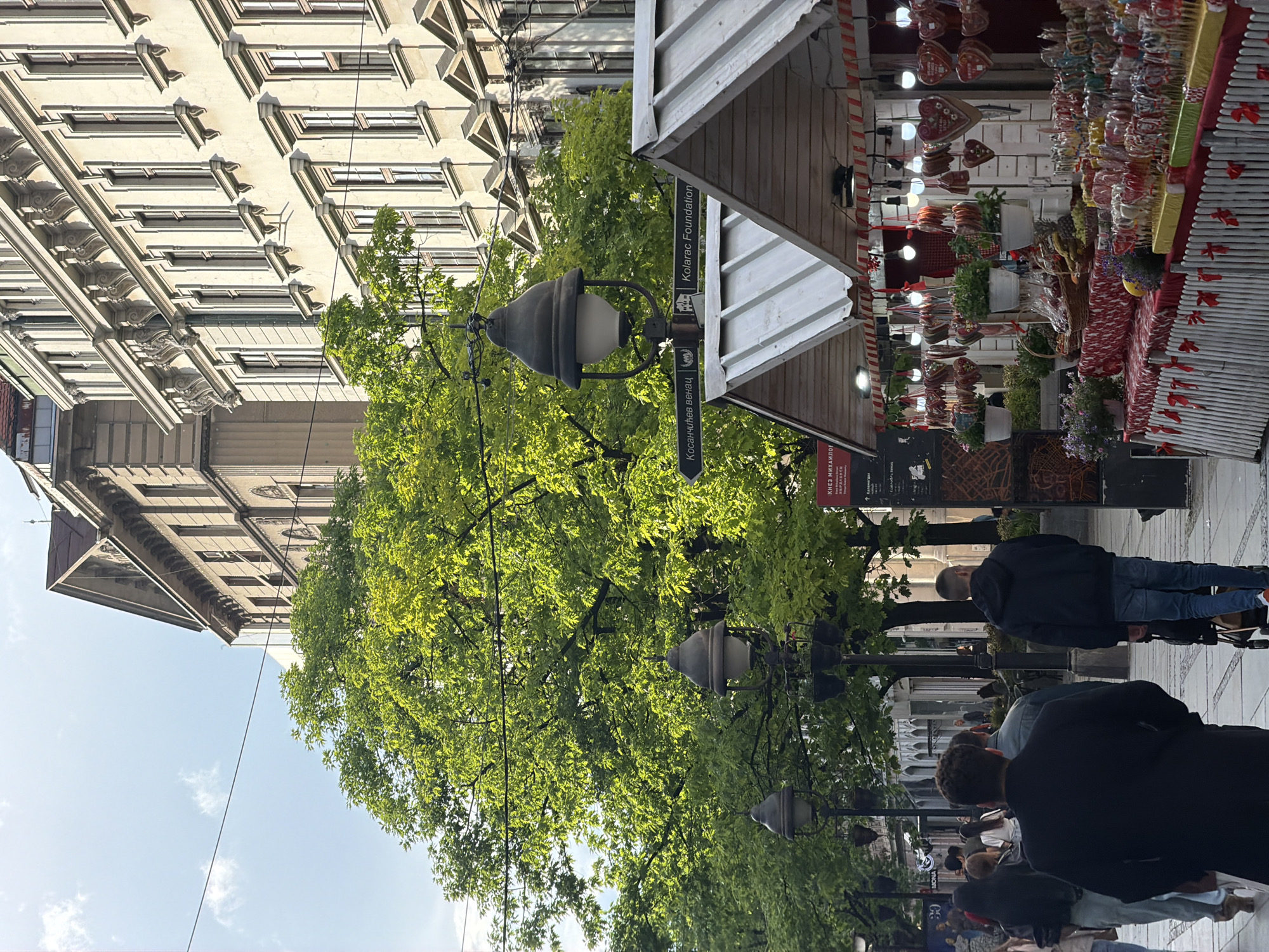 The image captures a street scene in Belgrade, Serbia, featuring a pedestrian walkway lined with trees and lamp posts. The scene includes several people walking along the walkway. A market stall is visible on the right, decorated with hanging heart-shaped decorations and other merchandise. Buildings line the street. Street signs on the lamp post point towards "Kosančićev venac" and "Kolarac Foundation".