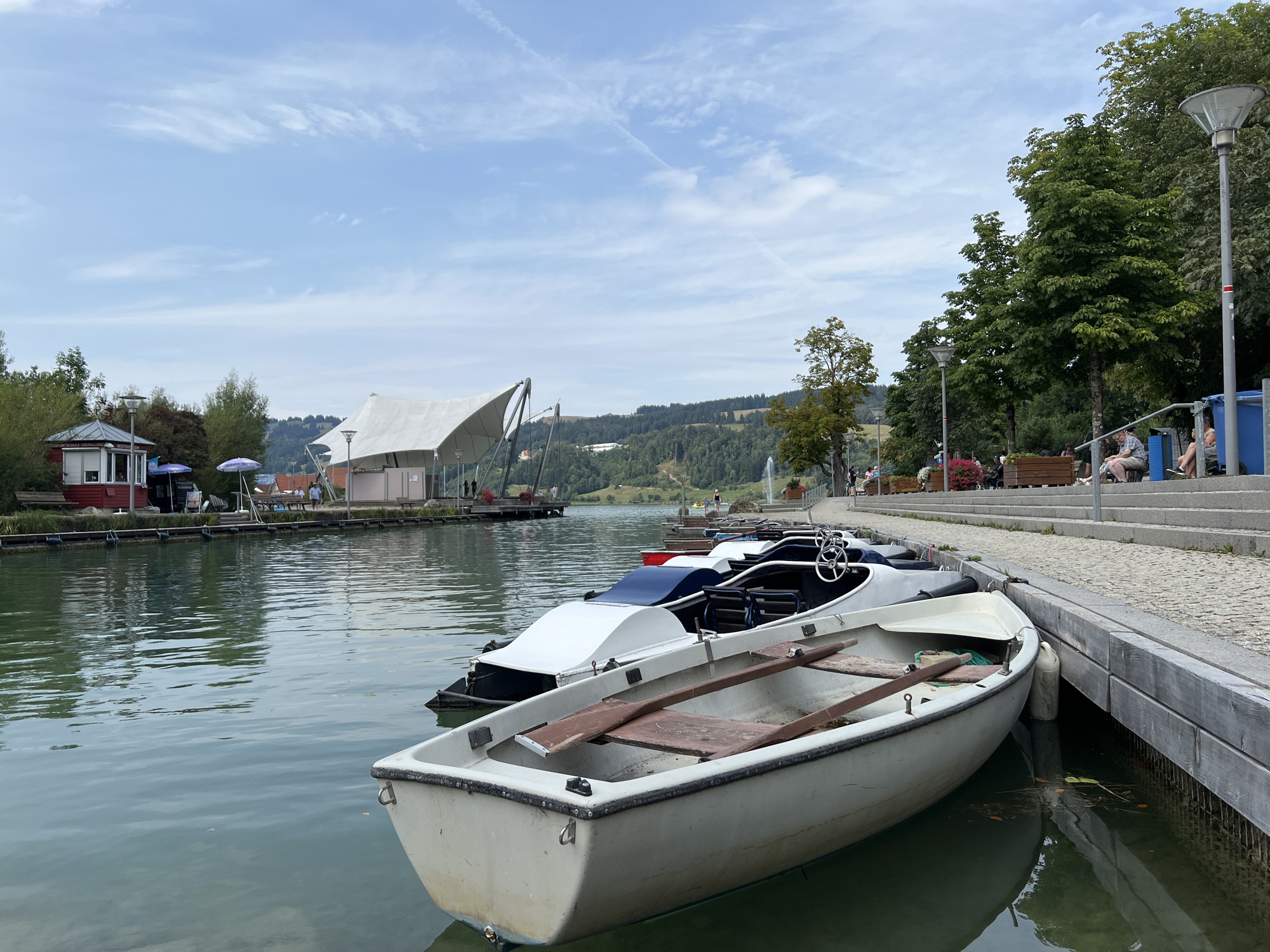 The image depicts a serene waterfront scene in Immenstadt im Allgäu, Germany, likely at the Immenstädter See. Several small boats, including rowboats and pedal boats, are moored alongside a wooden embankment. The water is calm and reflects the overcast sky and surrounding trees. In the background, a large white canopy structure, possibly a stage or pavilion, is visible, along with a small red building that might be a kiosk or rental booth. People are seen seated on benches and walking along the embankment, enjoying the pleasant weather. The landscape is characterized by lush green trees and rolling hills in the distance, typical of the Bavarian region. The overall impression is one of a tranquil recreational area.