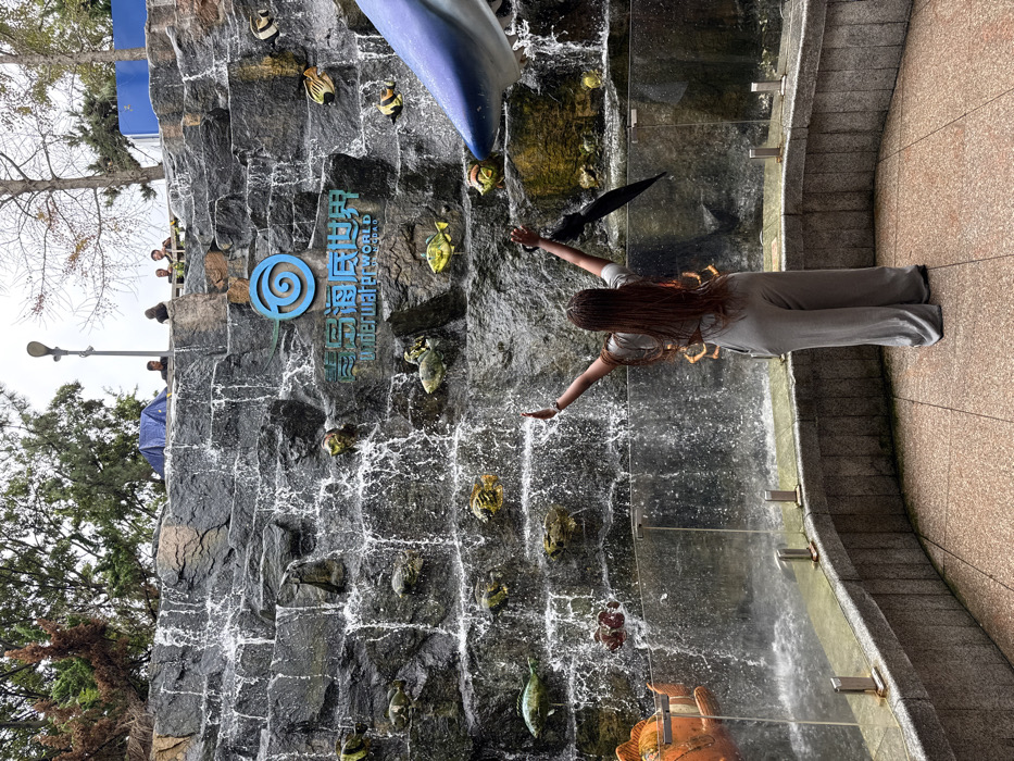 The image depicts a female individual standing with her back to the camera, arms raised in a "V" shape, facing a large artificial rock waterfall at the Qingdao Underwater World in Shinan District, Qingdao, China. She has long, reddish-brown braided hair and is wearing a light grey, loose-fitting two-piece outfit consisting of a crop top and wide-leg pants. A closed black umbrella is held in her left hand.
The multi-tiered waterfall cascades into a pool behind a curved, low concrete barrier topped with a transparent glass panel supported by metal stanchions. Embedded into the dark grey rock face of the waterfall are numerous colorful artificial fish models of various species. A prominent blue spiral logo and the white text "青島海底世界" (Qingdao Underwater World) are visible on the rock face above the waterfall's midpoint. At the lower left, a large orange clownfish model is partially visible, while a large blue and white shark head model is positioned on the lower right, extending into the frame.
Above the waterfall, on a raised platform or walkway, several indistinct individuals are visible, along with a blue tarp. The area behind and above the waterfall features green trees and some deciduous trees with bare branches, suggesting an outdoor environment. An outdoor lamppost is visible on the upper left. The sky is overcast and grey. The ground in the foreground is a light-colored tiled surface.