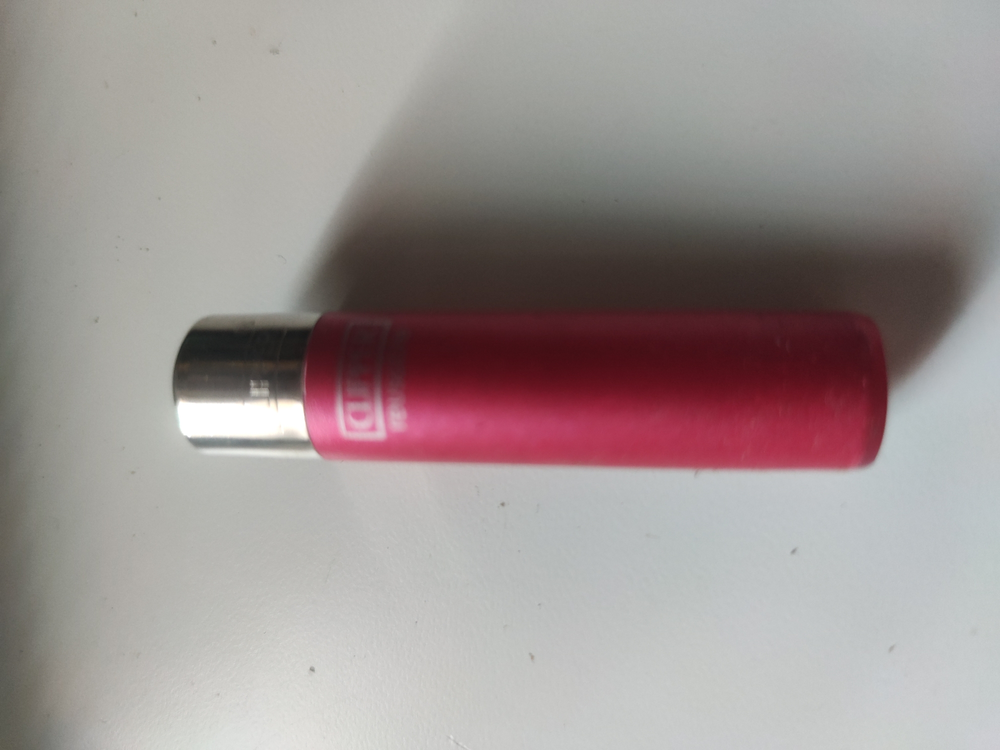 A pink Clipper lighter with a silver top is lying horizontally on a white surface. A shadow is cast behind the lighter. The city and environment of Unknown, Unknown are not ascertainable from the image.