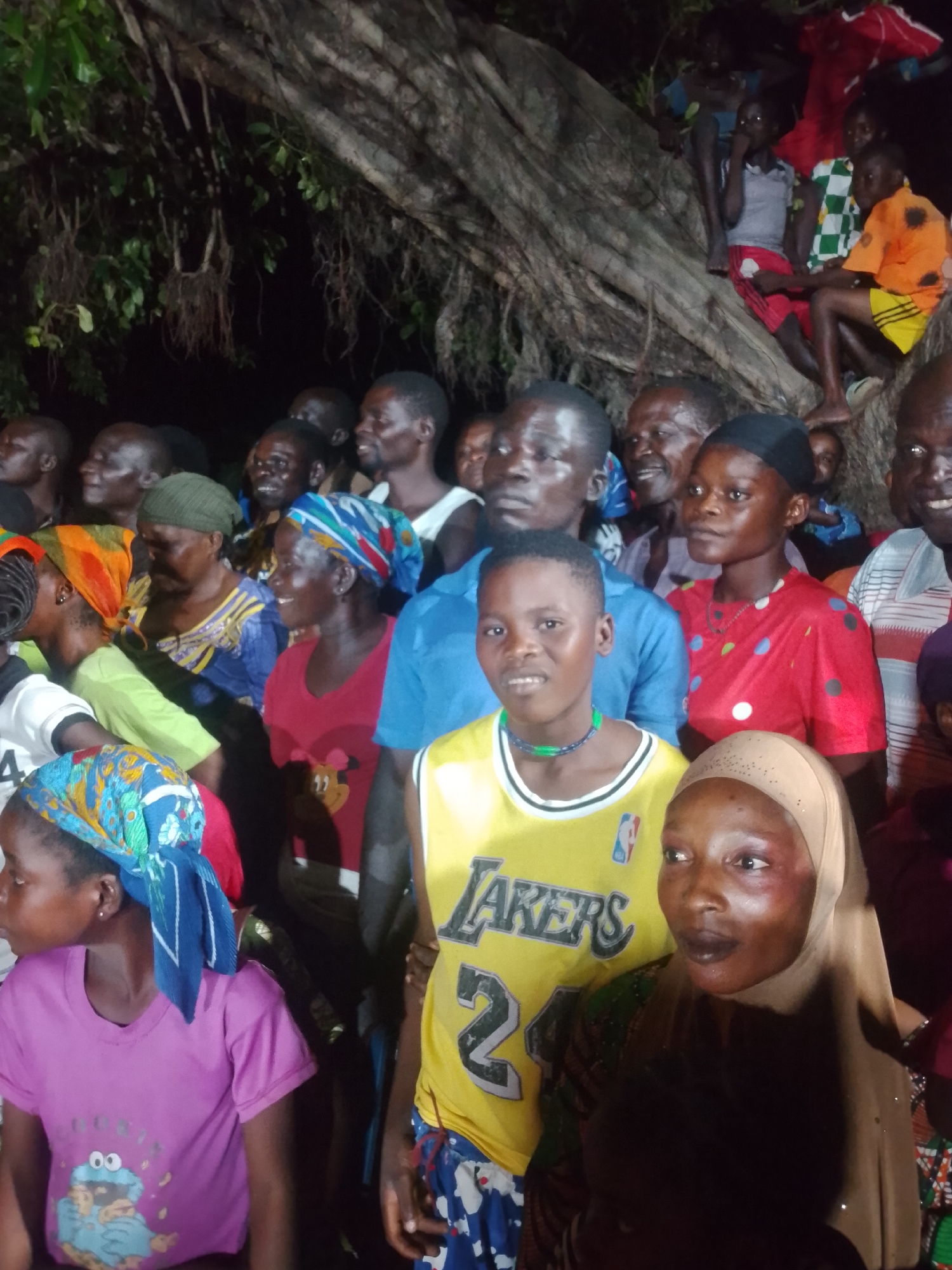 In the foreground of the image, a group of individuals, predominantly women and children, are gathered. A young person in a yellow basketball jersey with the word "LAKERS" and the number "24" is prominently positioned, looking towards the camera with a slight smile. To their right, a woman wearing a light brown hijab is visible, her head tilted to the side. To the left of the young person, a woman in a bright pink t-shirt with a cartoon character print is also facing left. Several other women are behind them, some wearing colorful headscarves and patterned clothing.
In the mid-ground, more people are visible, including men and women. Some individuals are looking in different directions, suggesting they are engaged in conversation or observing something outside the frame. A man in a blue shirt is partially visible.
In the background, a large tree with thick branches and aerial roots dominates the scene. Some individuals are seated on the branches of the tree, observing the gathering below. The lighting suggests the scene is taking place in the evening or at night, with artificial light sources illuminating the area. The environment appears to be outdoors, possibly a park or a communal gathering space. The overall mood seems to be one of community engagement or a special event.