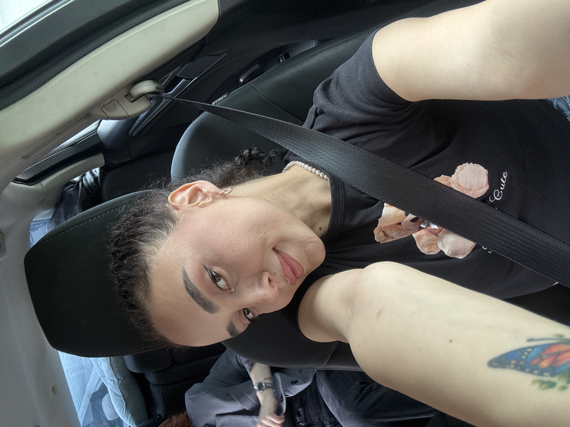 A woman is inside a vehicle, wearing a seatbelt and a black t-shirt with a bear design. She also wears a pearl necklace and has a tattoo on her arm. The vehicle appears to be in motion, given the blurred background and location is unknown.