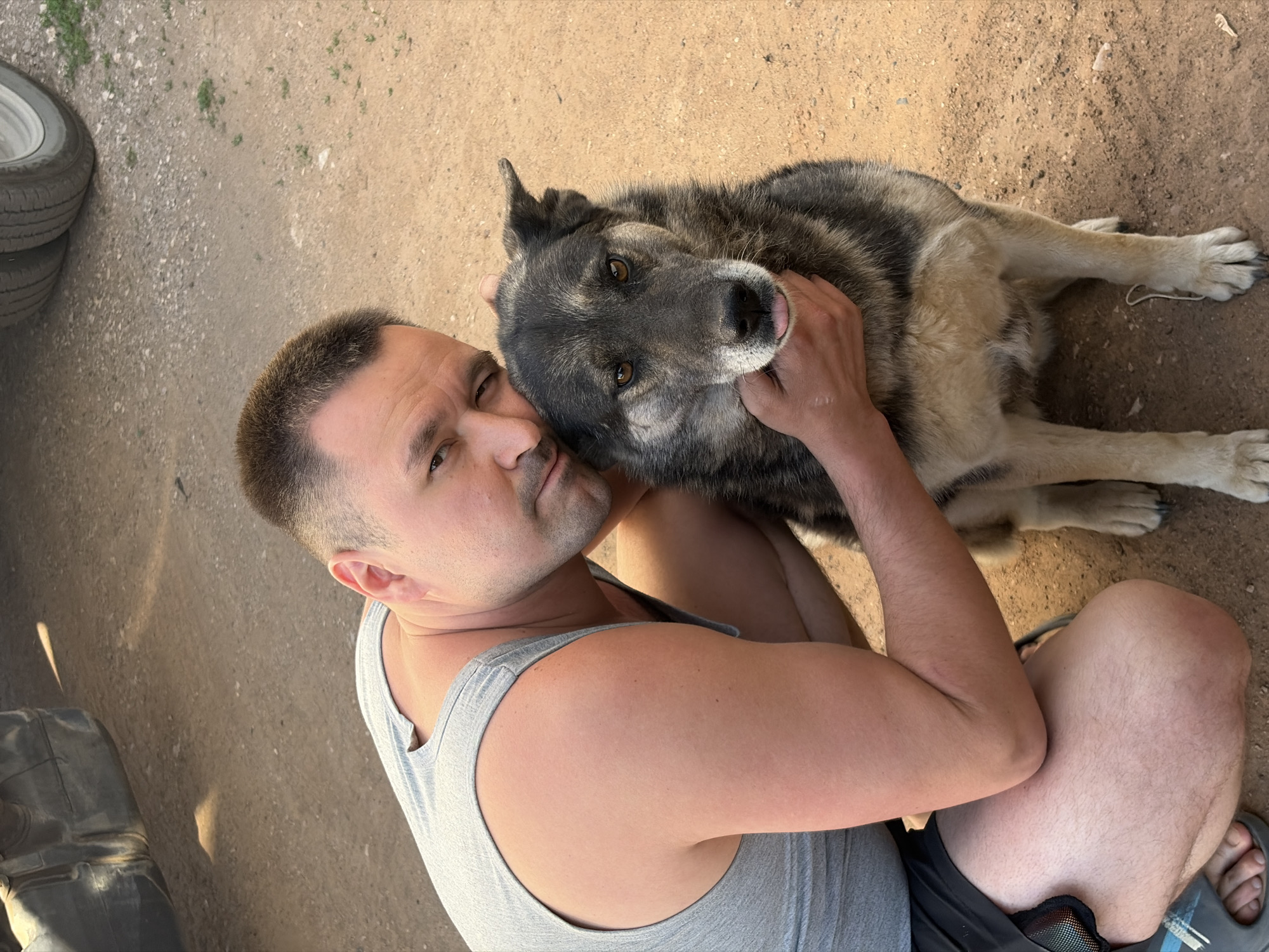 A man in a gray tank top is sitting cross-legged on dirt ground, embracing a large, gray and tan dog. The dog is lying down with its head resting on the man's chest. The man is looking at the camera with a slight smile. A stack of tires is visible in the background on the left. The location cannot be confirmed as Izhevsk, Russia based on the visual information in the image.