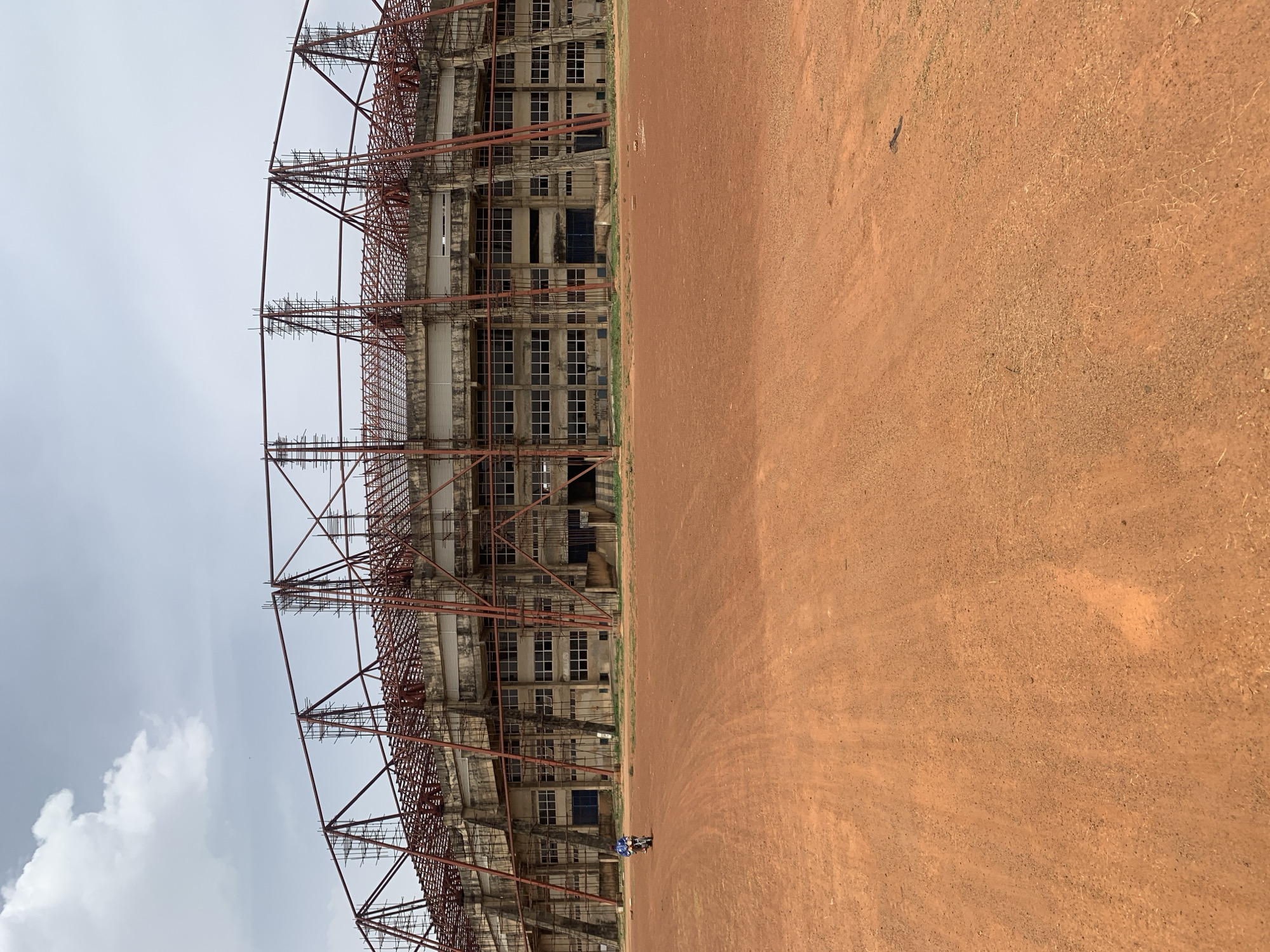 The image shows a large, unfinished concrete stadium structure with extensive metal scaffolding at an undetermined location in Rafin Par, Nigeria. The stadium's frame is visible through the scaffolding, showcasing multiple tiers of windows and support beams. The ground in front of the stadium is a reddish-brown dirt surface. A small figure is present on the ground near the stadium, possibly a person. The sky is overcast with grey clouds, though a patch of white cloud is visible. The overall scene suggests construction or renovation in progress.