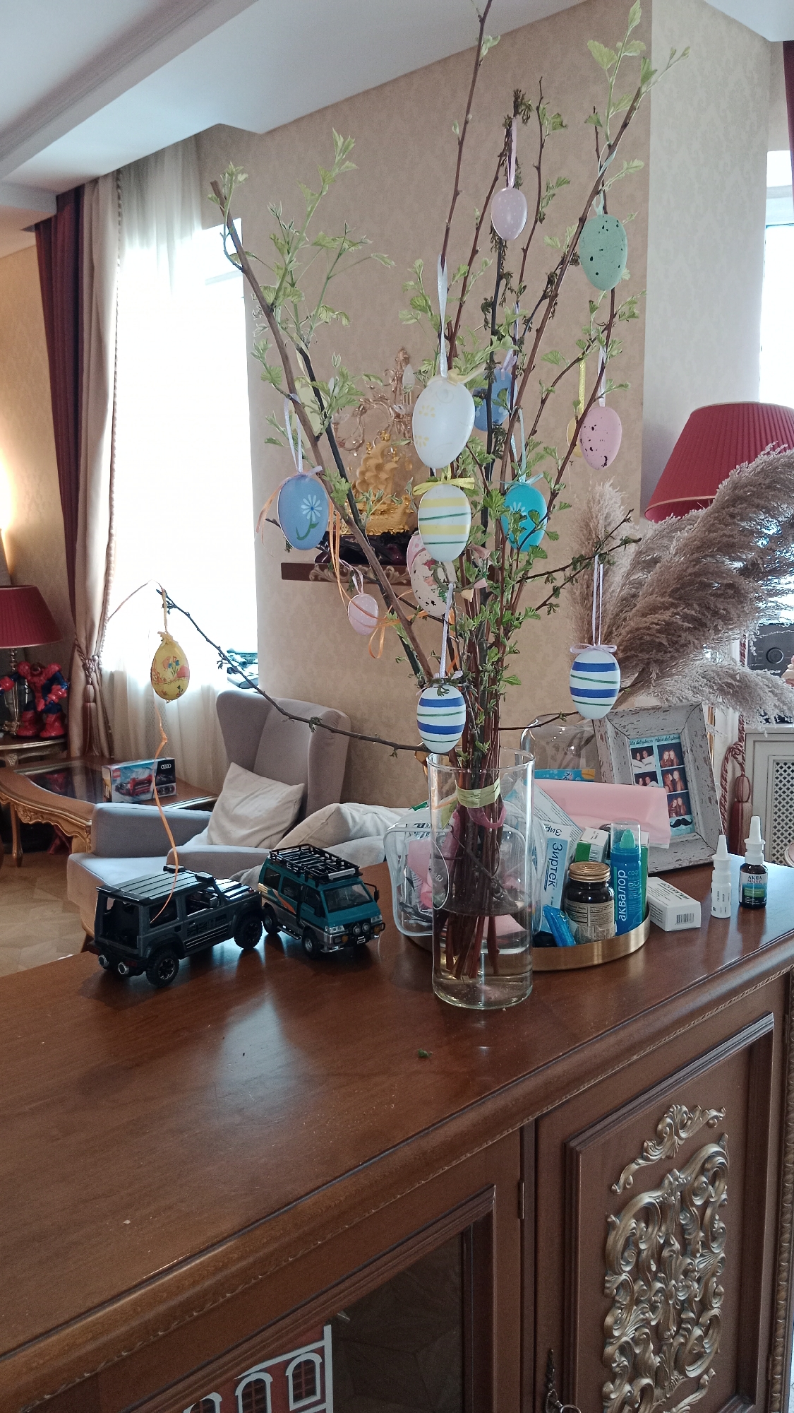 The image captures an interior scene likely within a home or apartment, identifiable by the presence of typical domestic furniture and decorations. The focal point of the scene is a dark wood cabinet on which an Easter decoration consisting of branches adorned with colorful Easter eggs is displayed in a glass vase. Adjacent to the vase are several medicinal items, including small bottles and boxes. Two toy vehicles, one black and one green, are positioned on the cabinet's surface. In the background, a softly lit living room area features a comfortable armchair, decorative pillows, and a side table with a table lamp. The room is illuminated by natural light coming through sheer curtains and a window. The style of the interior suggests a location in Moscow, Russia, based on common architectural and interior design trends. There is no visible people in this scene.