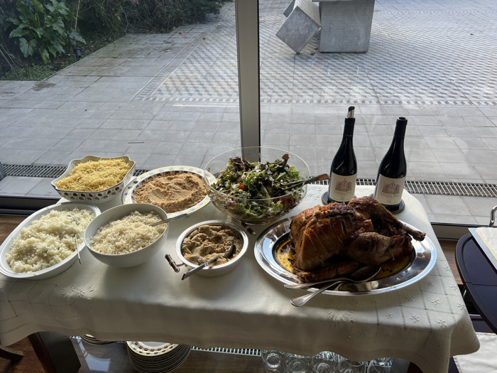 The image displays a buffet-style meal set indoors on a table covered with a white embroidered tablecloth. Visible dishes include a large roasted turkey on a silver platter, accompanied by two bottles of "Quinta do Poeira" wine (one with a pouring stopper) positioned behind it. To the left, a large glass bowl contains a mixed green salad. Other white serving dishes hold various side items: a bowl of yellow, thin, noodle-like strands; three separate servings of plain white rice in different containers; and two bowls containing a light brown, granular, or crumbly substance, resembling farofa. Serving utensils are present in most dishes. Below the table, a stack of patterned plates and several empty glasses are visible.
The scene is set against a large window or glass door, offering a view of an outdoor area. This outdoor space features a paved surface with a distinctive geometric, checkered pattern. To the left, green foliage is visible. To the right, a large, abstract sculpture composed of multiple grey, cube-like concrete elements stands on the paved ground. The location is identified as a building in Porto, Portugal. No individuals are present in the scene.