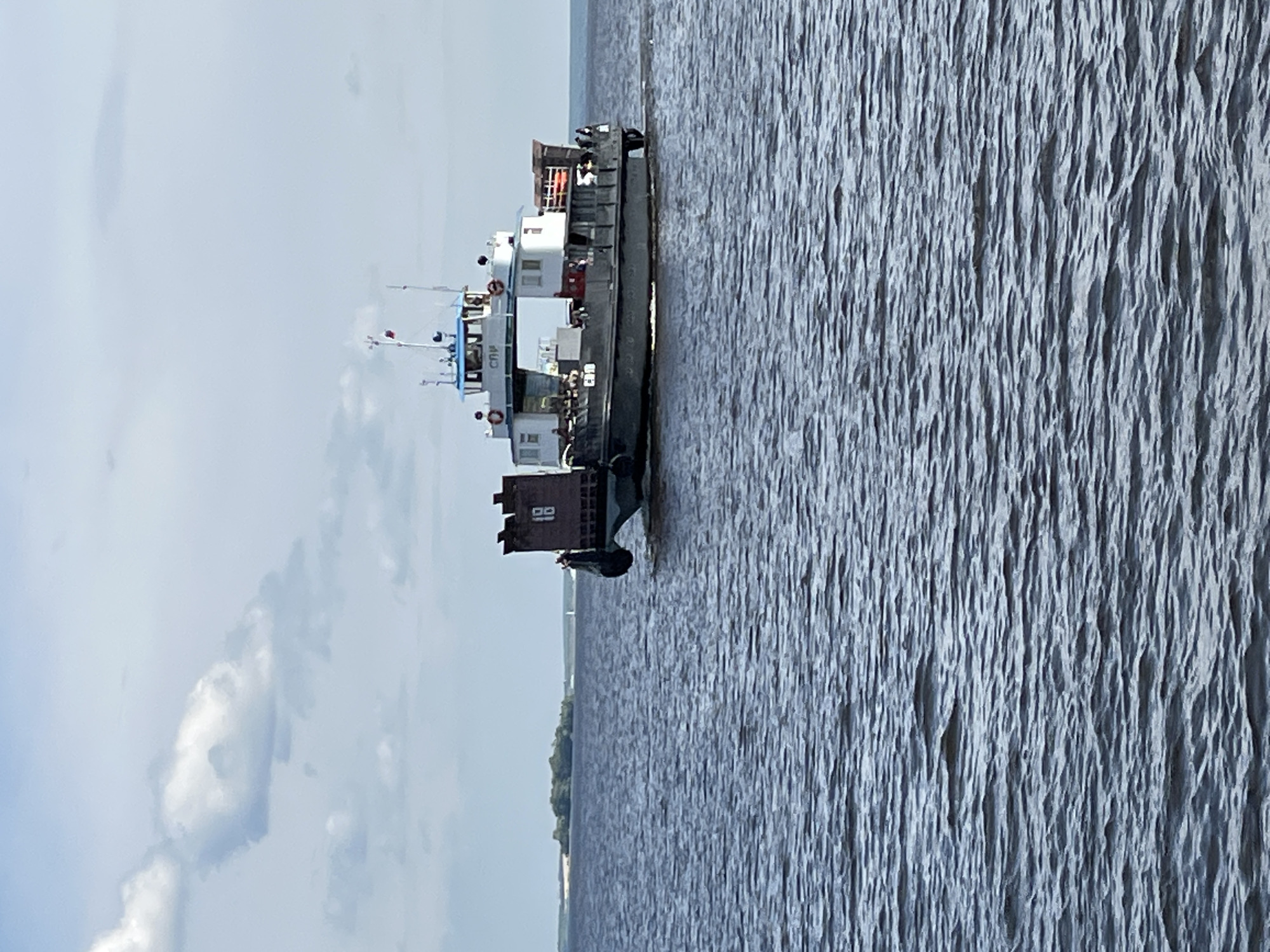 The scene depicts a ferry vessel navigating a wide body of water under a partially cloudy sky in Kazan, Russia, likely on the Volga River or Kuybyshev Reservoir.
The primary subject is a multi-deck, grey and white ferry, positioned in the mid-ground and moving from the left side of the frame towards the right. The vessel features a dark grey lower hull and a lighter grey and white superstructure. A prominent dark grey ramp or gate, marked with the white numeral "18," is visible on the bow. The letters "СВП" are painted in dark Cyrillic script on the white side of the main superstructure, below the pilothouse. The upper deck includes a distinctive blue and white pilothouse, topped with antennas and a small flag, which appears to be the Russian tricolor (white, blue, red). Several individuals are discernible on the open decks of the ferry, appearing to be passengers or crew members. The ferry is actively moving, creating a visible wake behind its stern.
The water surface is dark blue-grey with moderate ripples covering the foreground and mid-ground. In the distant background to the left, a low-lying landmass or island covered with green vegetation and a lighter sandy shore is visible. The sky above is a pale blue with scattered white cumulus clouds. A very small, indistinct white object, possibly a sailboat or buoy, is faintly visible on the distant horizon line to the far right.