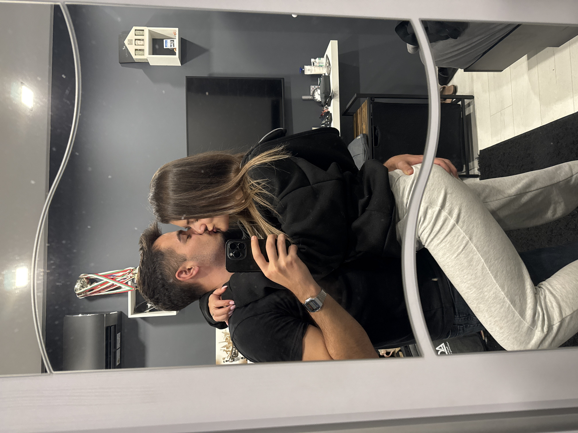 A couple kisses in a mirror selfie. The man wears a black t-shirt and a silver watch, and holds the phone with his right hand. The woman wears a black hoodie and grey sweatpants.
The room's decor includes a wall-mounted model house, a framed trophy on a shelf, a white shelf with various items, a TV, and a black table. The flooring appears to be wooden with a black rug.
There is no information to determine the specific location within Sofia, Bulgaria.
