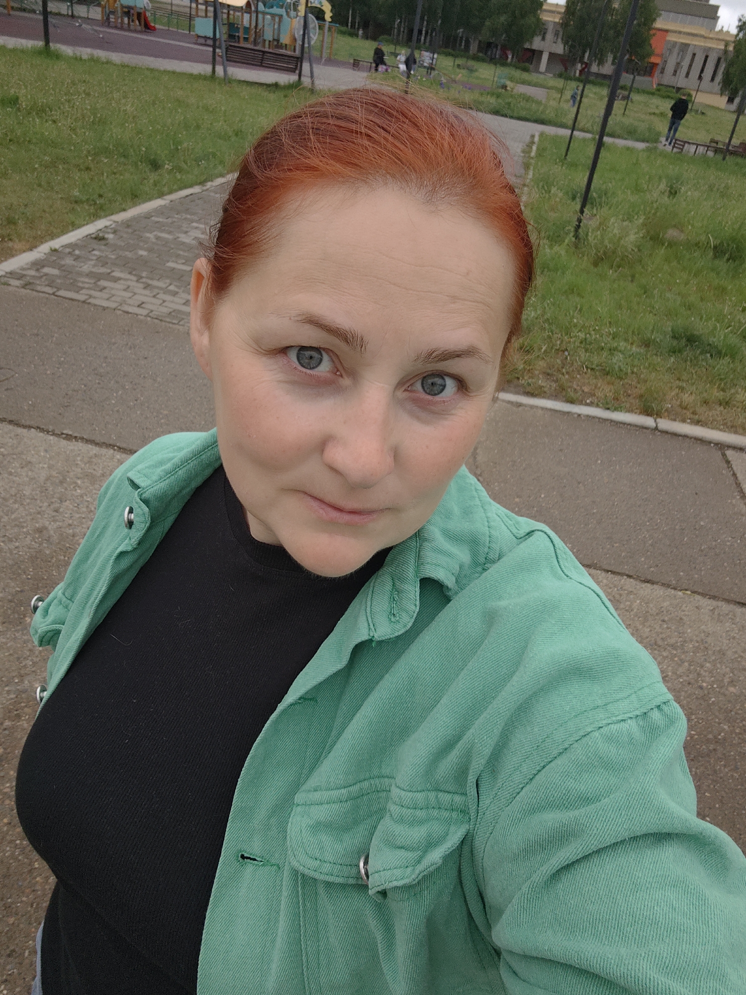 A medium close-up, eye-level selfie shows a woman with red hair and blue eyes looking directly at the camera. She wears a black shirt underneath an unbuttoned mint green denim jacket. The background features a paved path leading to a grassy area and a playground with colorful equipment. Trees and a building are visible further in the background, suggesting a park setting. In the distance, there are a few people walking. The setting appears to be a city park in Марковский, Russia.
