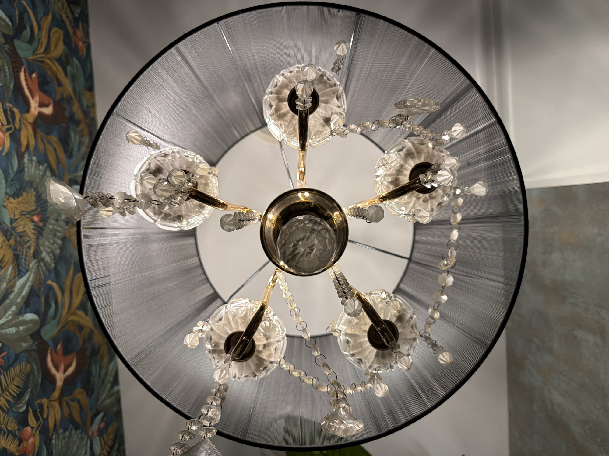 View of a chandelier with five crystal lights and a large, black drum shade, photographed looking directly up. The chandelier's central metal structure is brass-colored. The surrounding environment includes a section of a wall with floral wallpaper to the left, and a gray wall to the right. The ceiling is white. There is no evidence suggesting the location is in Chaikovsky, Russia.