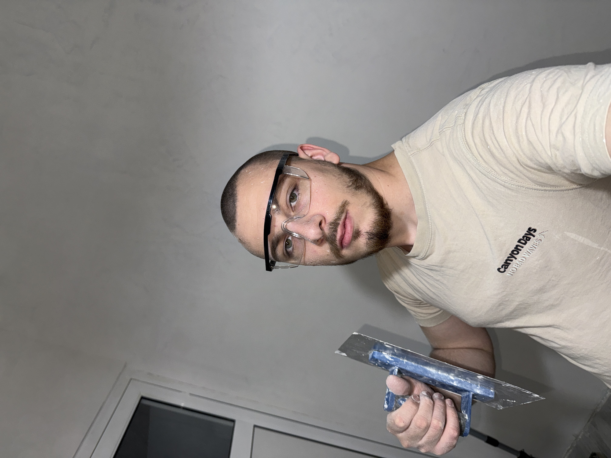 A man with a beard and wearing safety glasses holds a putty knife. He is wearing a light-colored t-shirt with the words "Canyon Days NO BAD WAVES" printed on it. The background appears to be a white wall and ceiling, with a window visible at the bottom left. It is not possible to verify the location as Stara Zagora, Bulgaria from this image.
