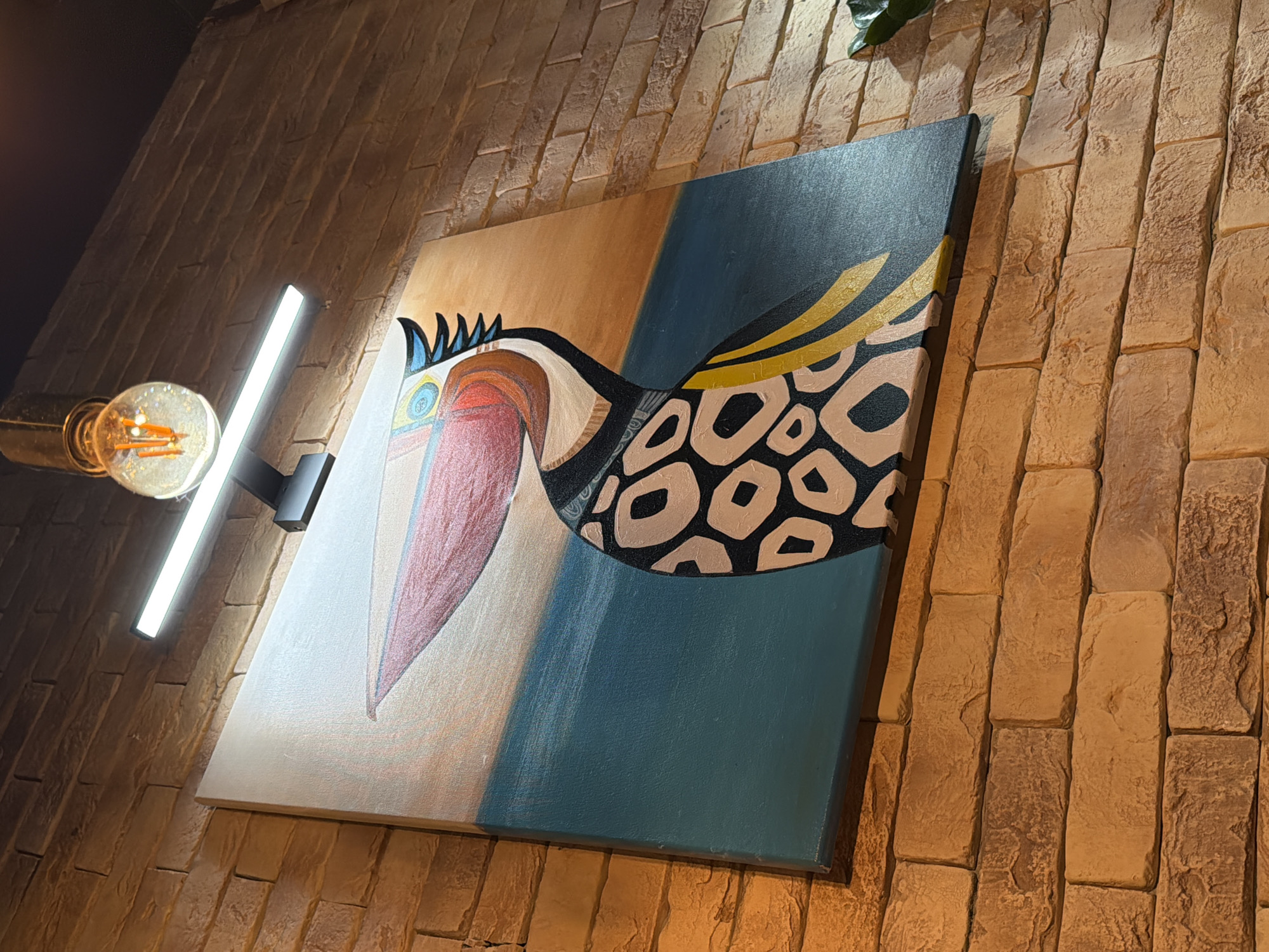 A brightly lit close-up shows a painting of a bird in a room with tan brick walls. The painting is of an abstract bird with a large red beak, and a wing made of leopard-like circles. The painting has three color blocks, a tan color where the head is, a white block to the left of the beak, and a blue background for the wing. A vertical light is visible on the left side of the painting. The location is not visible in the image. Therefore, the location in the city of Chaikovsky, Russia, is not available.