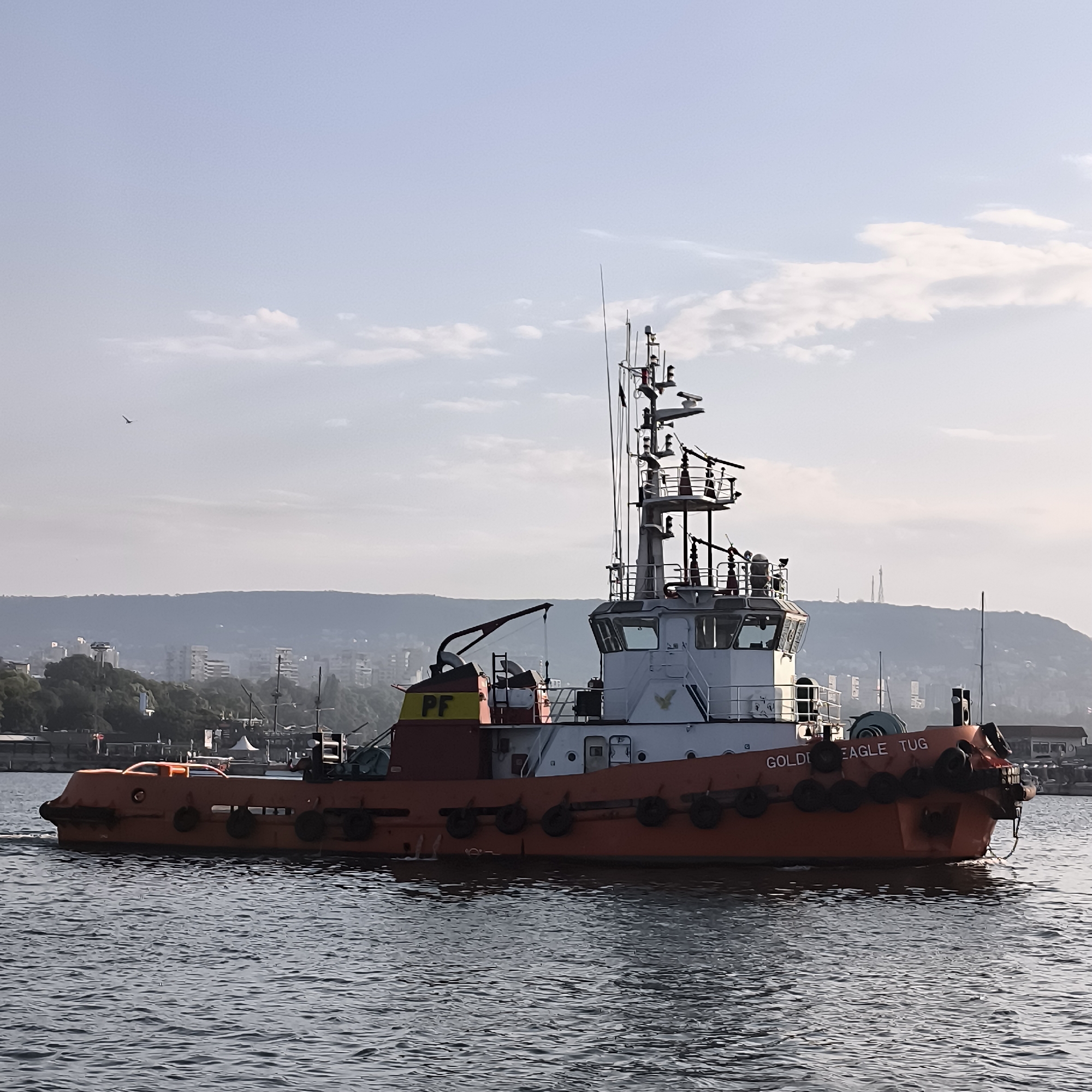The image depicts a tugboat named "GOLDEN EAGLE TUG" sailing on a body of water, likely the Varna Bay in Varna, Bulgaria. The tugboat is predominantly orange with white and red accents. It has a multi-level superstructure with various antennas and equipment, suggesting operational capability. The vessel is in motion, indicated by the slight wake it is creating in the water.
In the background, a hazy cityscape of Varna is visible, nestled against a backdrop of rolling hills. Numerous buildings, some appearing to be residential or commercial, can be discerned. The sky is partly cloudy with a light blue hue, suggesting it is either early morning or late afternoon. The overall atmosphere is calm and industrial, typical of a port city. There are no people clearly visible on the tugboat or in the surrounding environment. The presence of the city and its hilly terrain strongly points to the location being within the vicinity of Varna's port facilities.