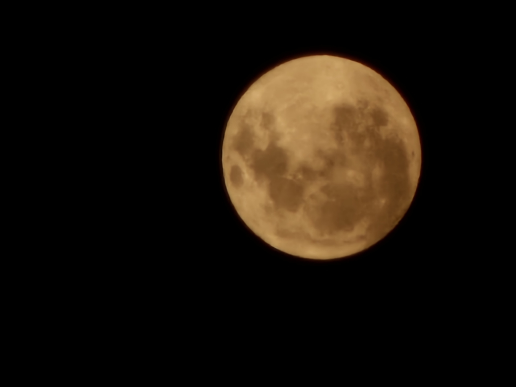 A close-up, side-view, vertical shot shows a full moon against a black, starless sky. The moon is a warm, golden-orange color and appears to be partially obscured by clouds or atmospheric haze, giving it a textured and slightly blurred appearance. The surface details of the moon, such as craters and maria, are visible. The image was taken in Denpasar, Indonesia.