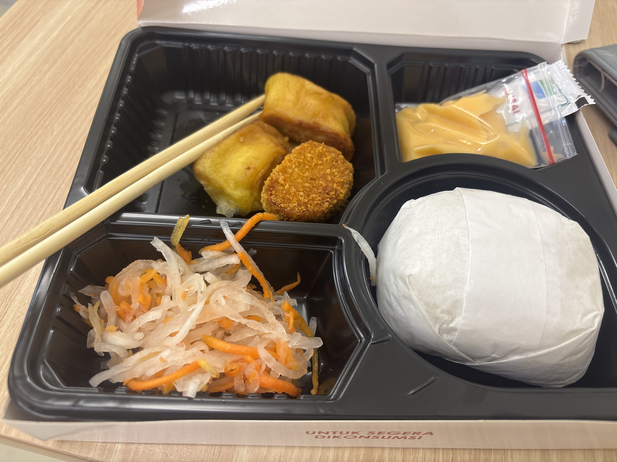 A close-up, eye-level shot shows a disposable black plastic bento box filled with food, resting on a light brown wooden surface. The bento box has compartments. In the top left compartment, two wooden chopsticks lie diagonally across two golden-brown, rectangular fried items and a small, round, breaded item. To the right of the fried items, in a separate compartment, is a small, clear plastic bag filled with a yellow, creamy sauce. In the bottom left compartment, a mixture of shredded white and orange vegetables, possibly pickled radish and carrot, is visible. In the bottom right compartment, a round food item wrapped in white paper sits. The bento box is partially open, revealing a white lid or outer packaging. The wooden surface appears to be a table. Text is visible on the edge of the bento box's outer packaging in red and black letters. The lighting suggests indoor illumination. The scene depicts a meal prepared for consumption. This image was taken in East Jakarta, Indonesia.