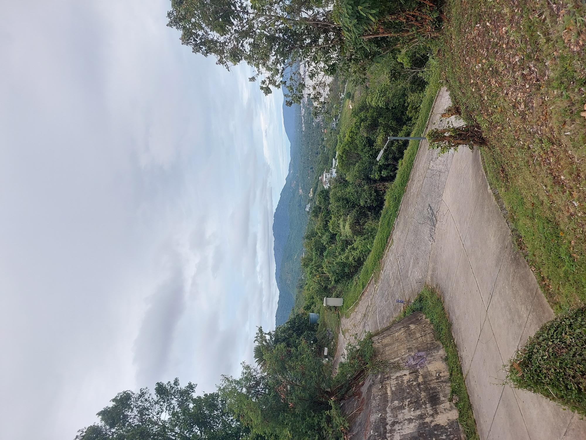 The image shows a paved road winding through lush green hills. The road is cracked and shows signs of wear. Trees and dense vegetation cover the slopes, with a few houses visible in the distance. The sky is overcast with gray clouds. The overall scene suggests a rural or semi-rural area with a natural landscape. The location is identified as Ko Samui District, Thailand, which is known for its tropical scenery and hilly terrain. There are no people or animals visible in the image, and no discernible actions or interactions taking place. The environment is dominated by natural elements like trees and hills, with the road and some utility structures being the only man-made elements.